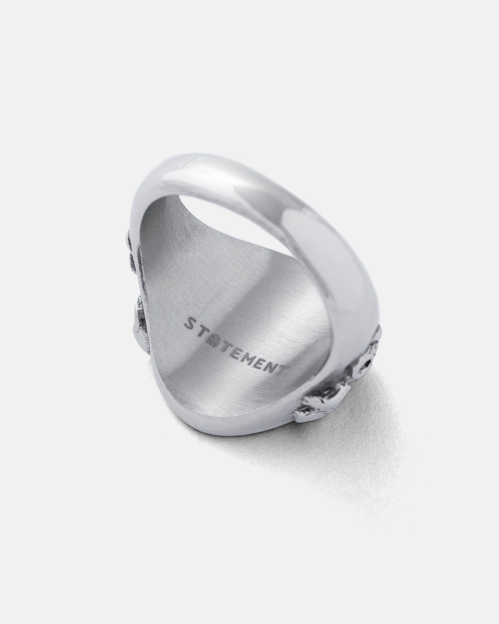 The Graveyard Signet Ring is a silver stainless steel ring with a polished, engraved interior reading STATEMENT and a textured outer edge for a modern spin on classic graveyard jewelry. Shown against a plain light background.