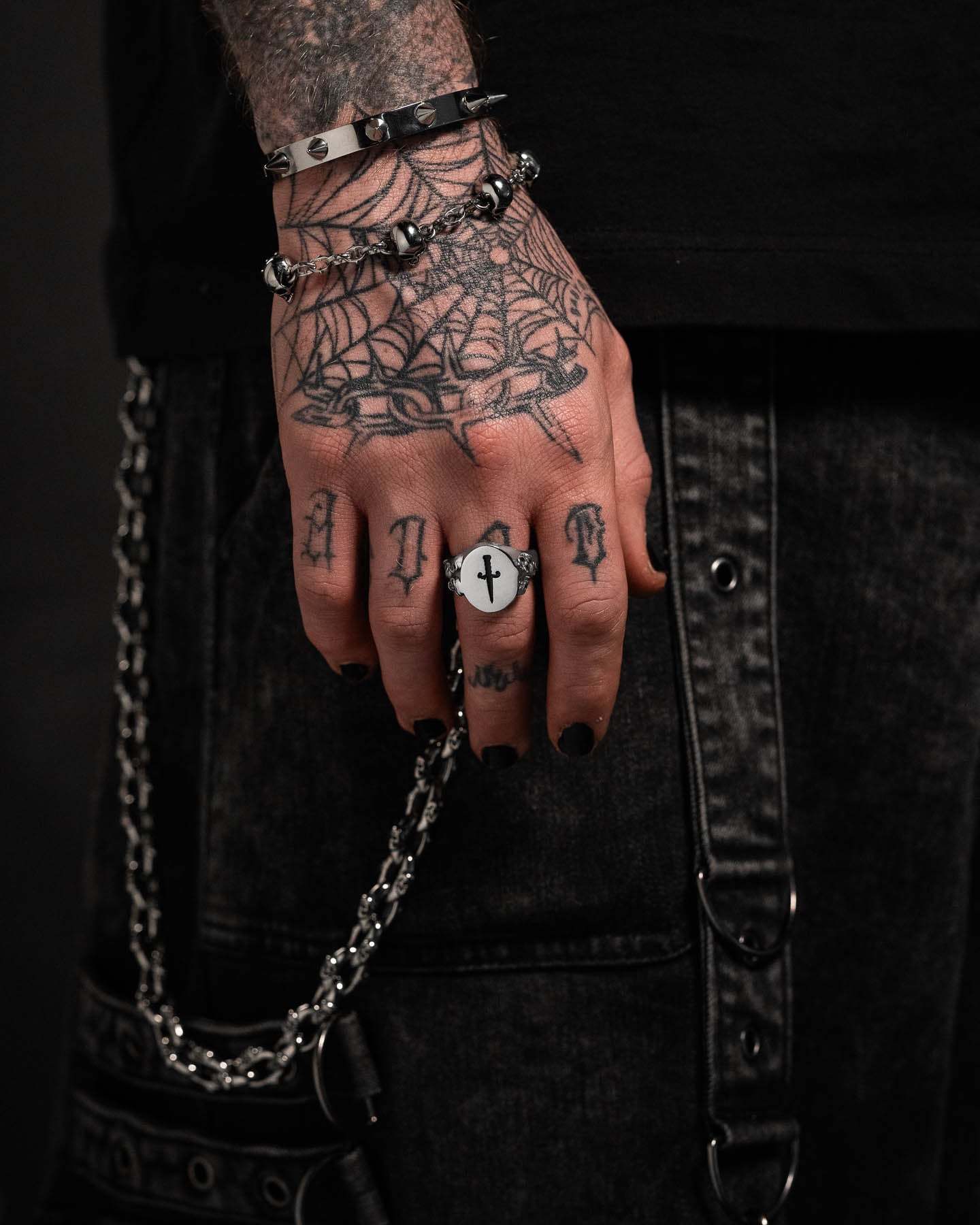 A tattooed hand with black nail polish, wearing the Graveyard Signet Ring and silver bracelets, rests against dark denim pants with metal chains. Visible tattoos include a spider web and letters on the fingers.
