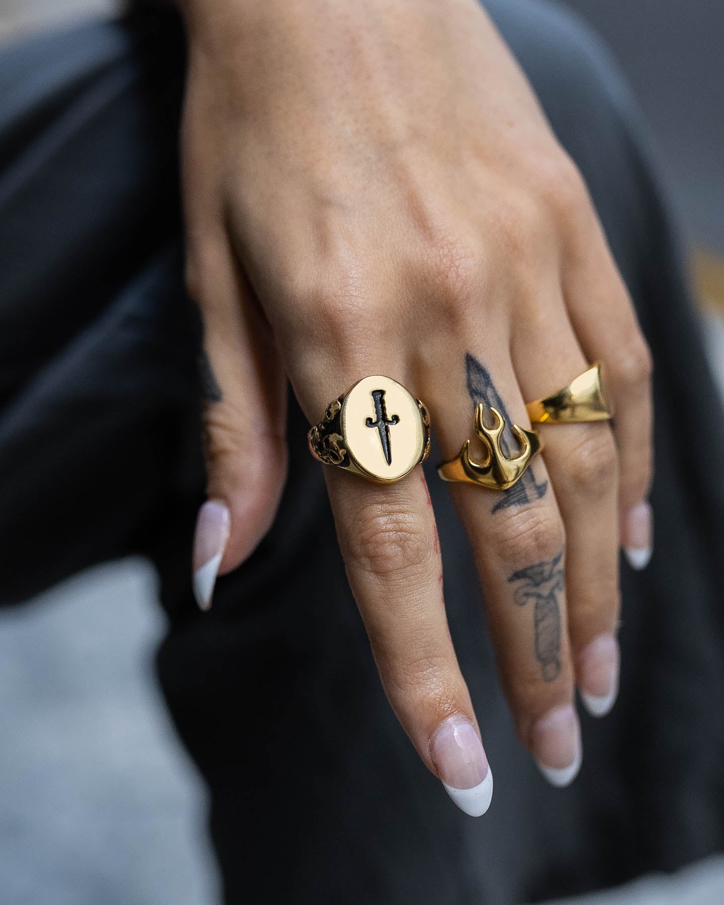A hand with long, almond-shaped nails wears three gold rings, including the Graveyard Signet Ring (Gold) and another with a sword design. A sword tattoo is visible on the middle finger as the hand rests on a dark surface.