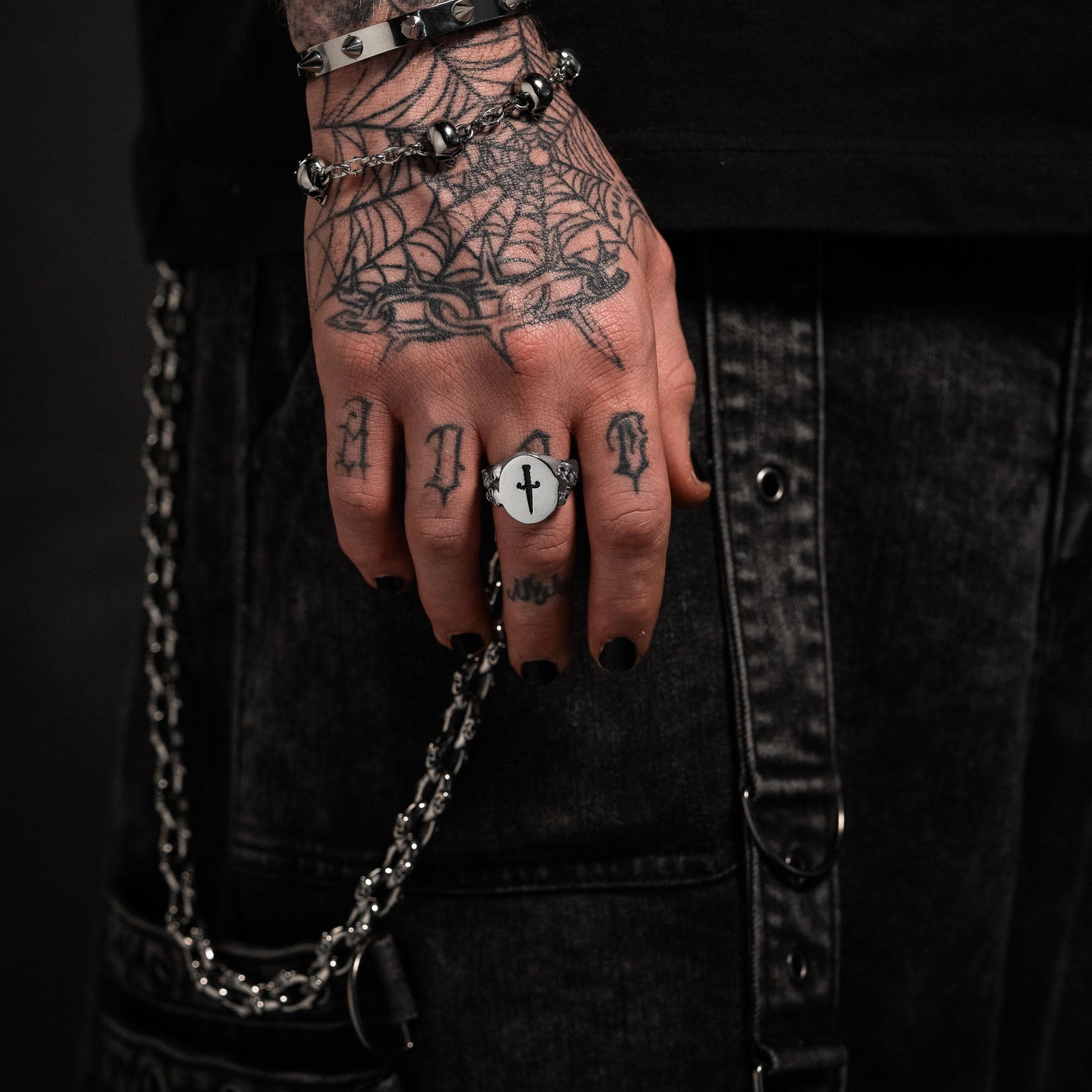 Hand with tattoos and a mens signet ring, wearing a black shirt on a dark background