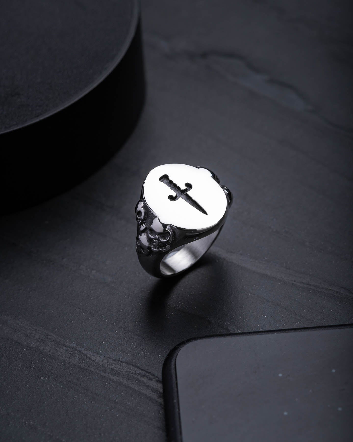 The Graveyard Signet Ring features a silver, flat oval top engraved with a black sword design. Small skull details decorate the sides, giving this signet ring an edgy and bold look.