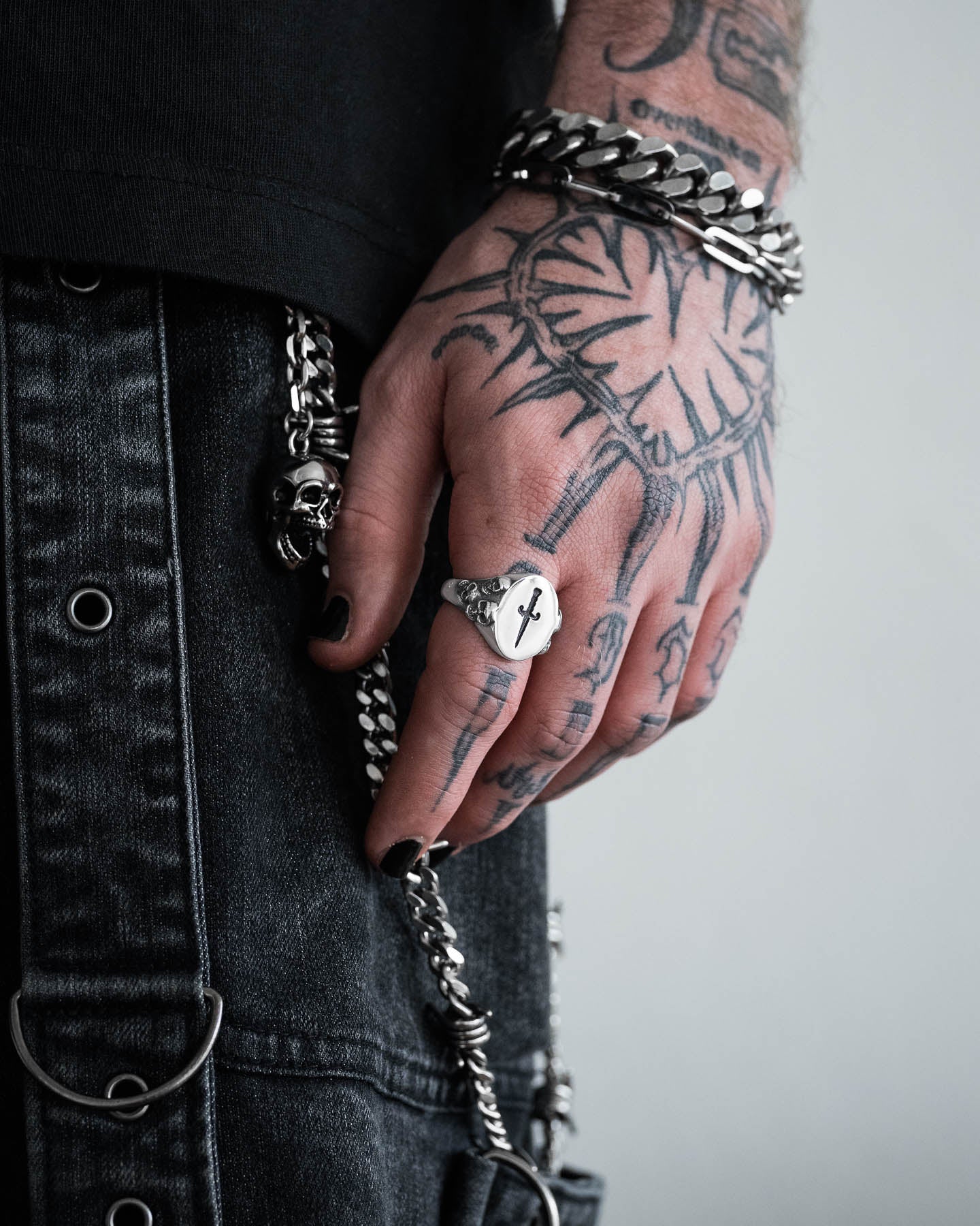 A tattooed hand with black-painted nails wears the Graveyard Signet Ring and a skull bracelet, paired with a chain bracelet. Dressed in black, they complete their style with metal accessories and decorative chains on their pants.