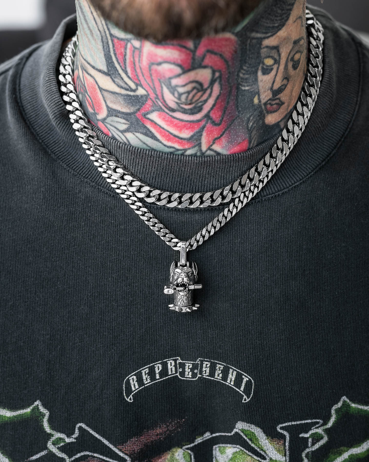 A close-up of a neck with colorful floral and portrait tattoos, wearing two silver chains—one plain and one bold street necklace featuring "The Guard Dog" Pendant—over a black graphic REPRESENT t-shirt.