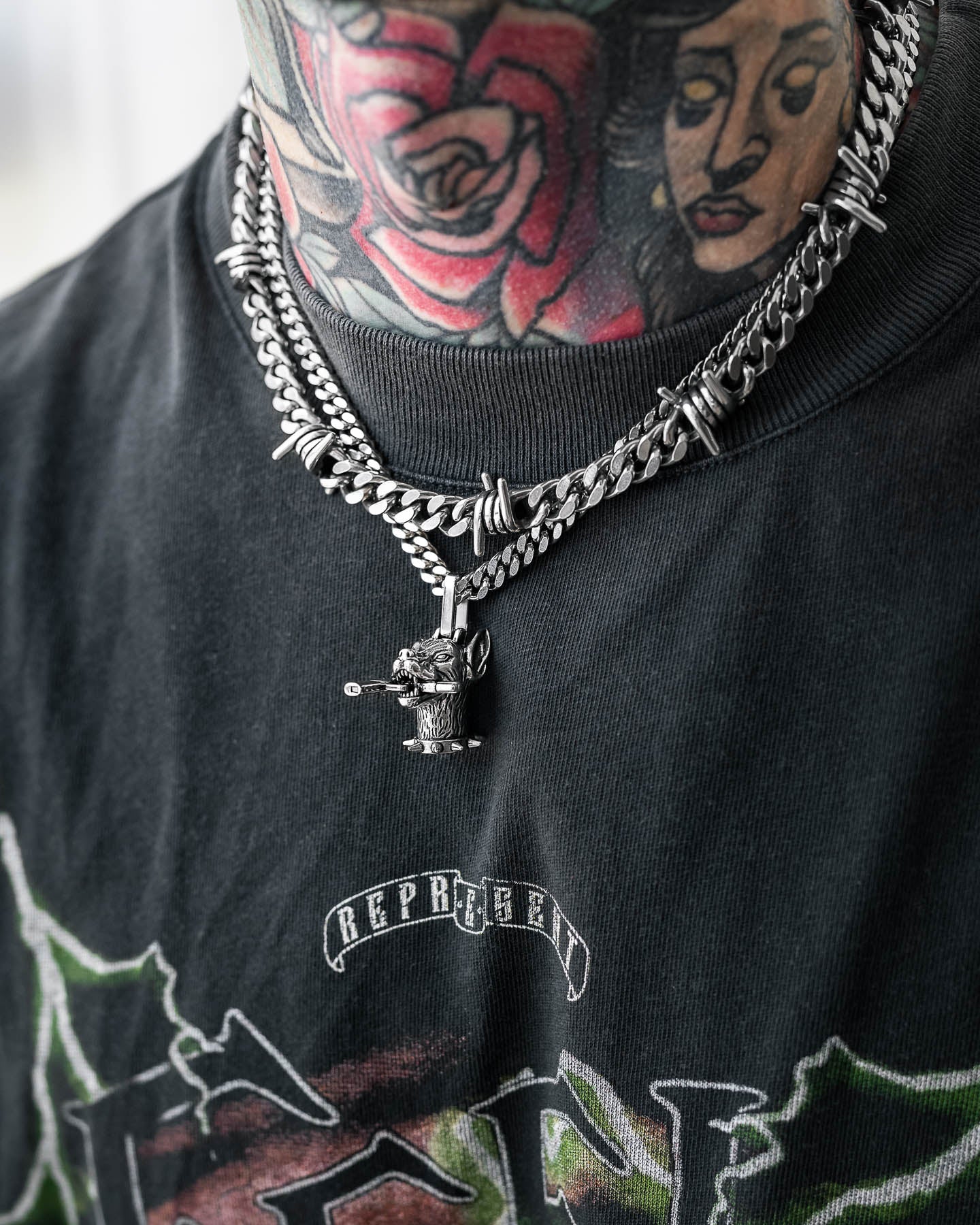A close-up shows a person with colorful rose and woman's face neck tattoos wearing "The Guard Dog" Set layered silver chains, featuring a barbed wire Cuban necklace with a bear pendant, paired with a black graphic T-shirt.