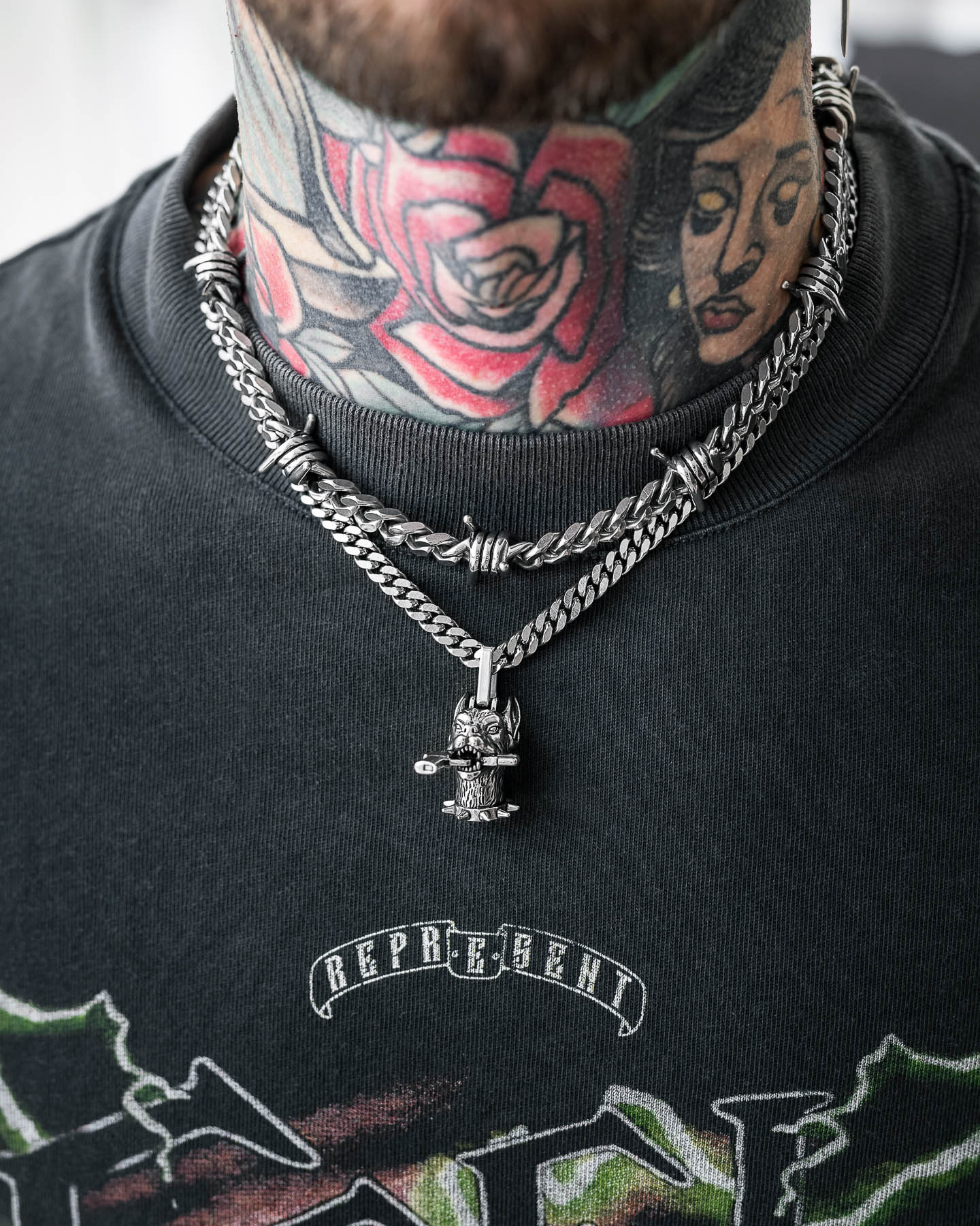 A person with colorful neck tattoos—featuring a large rose and a woman's face—wears "The Guard Dog" Set, two silver chains including a barbed wire Cuban necklace. They're in a black REPRESENT graphic tee.