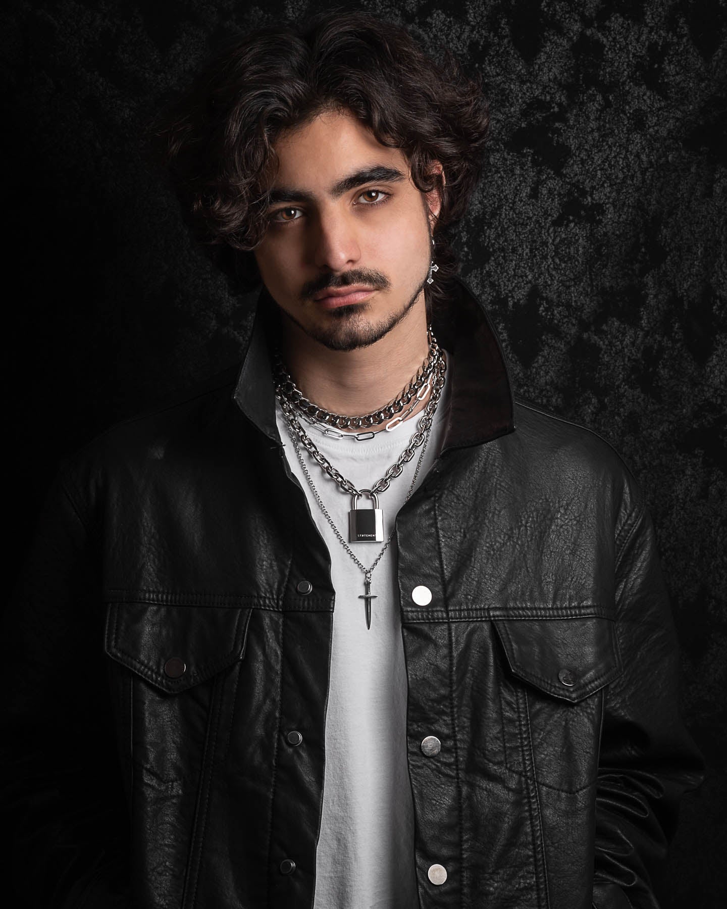 A young man with wavy dark hair and facial hair wears a black leather jacket over a white shirt, showing off the Handle With Care 2.0 Stack—a layered silver men’s necklace—while posing against a textured dark background with a neutral expression.