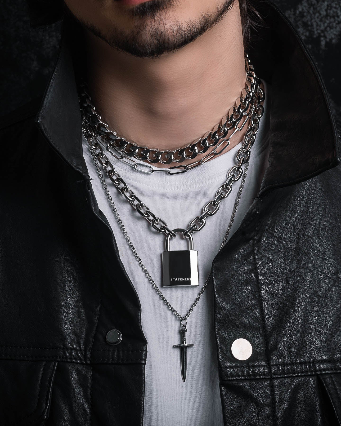 A person wears the Handle With Care 2.0 Stack—layered silver chain link necklaces with a padlock and dagger pendant—over a white shirt and black leather jacket, showing only the lower half of their face.