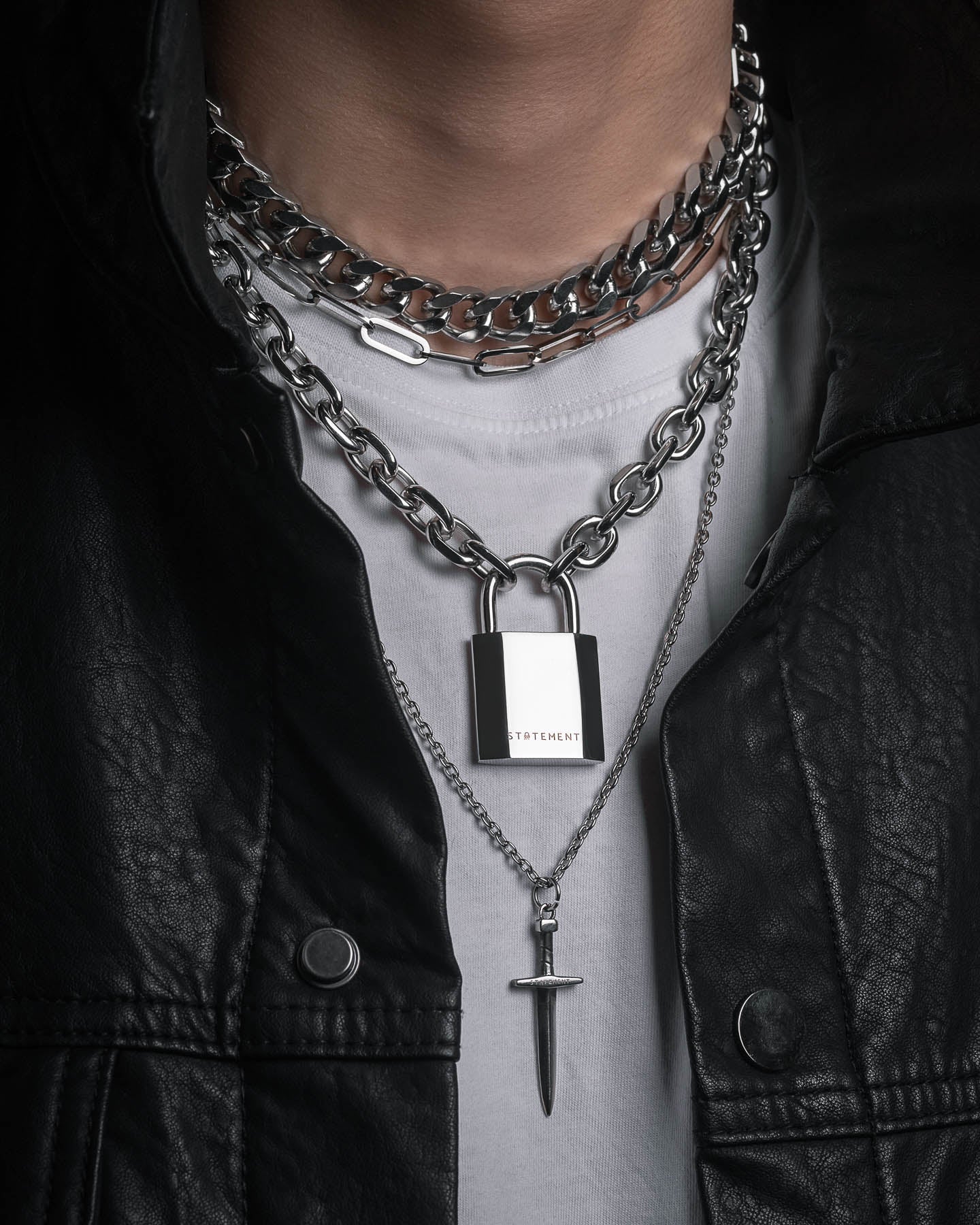 A person in a black leather jacket and white shirt, accessorized with the Handle With Care 2.0 Stack: bold men’s layered silver chains featuring padlock and dagger pendants, chunky chain links, and striking jewelry pieces.