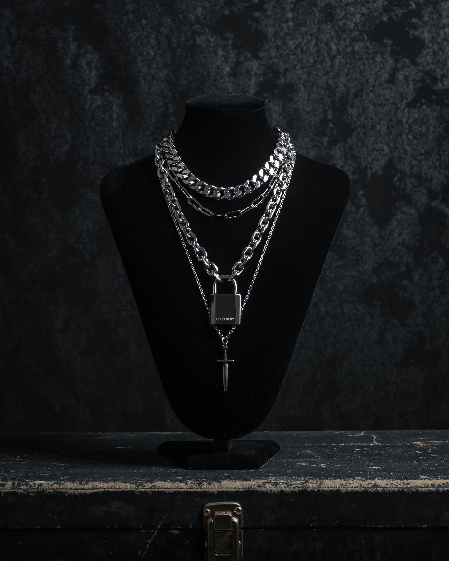 The Handle With Care 2.0 Stack, a men's silver chain necklace set featuring chunky links with lock and dagger pendants, is displayed on a black velvet jewelry bust against a dark textured background.