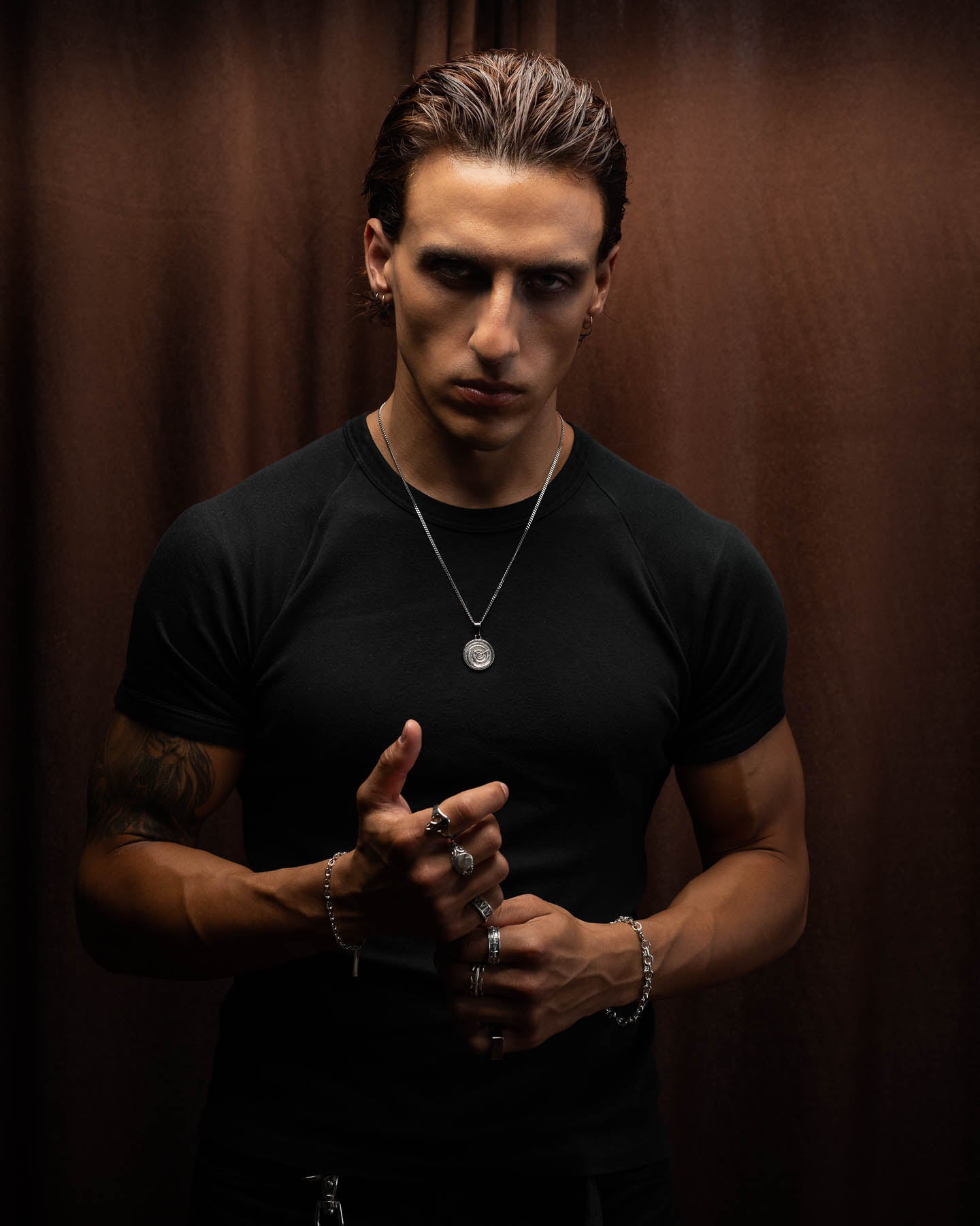 A man with slicked-back hair stands against a brown curtain, wearing a black shirt, silver jewelry, and rings. He holds the Infinite Clock Pendant Necklace—a Dali-inspired 316L stainless steel spiral clock pendant—between his hands.