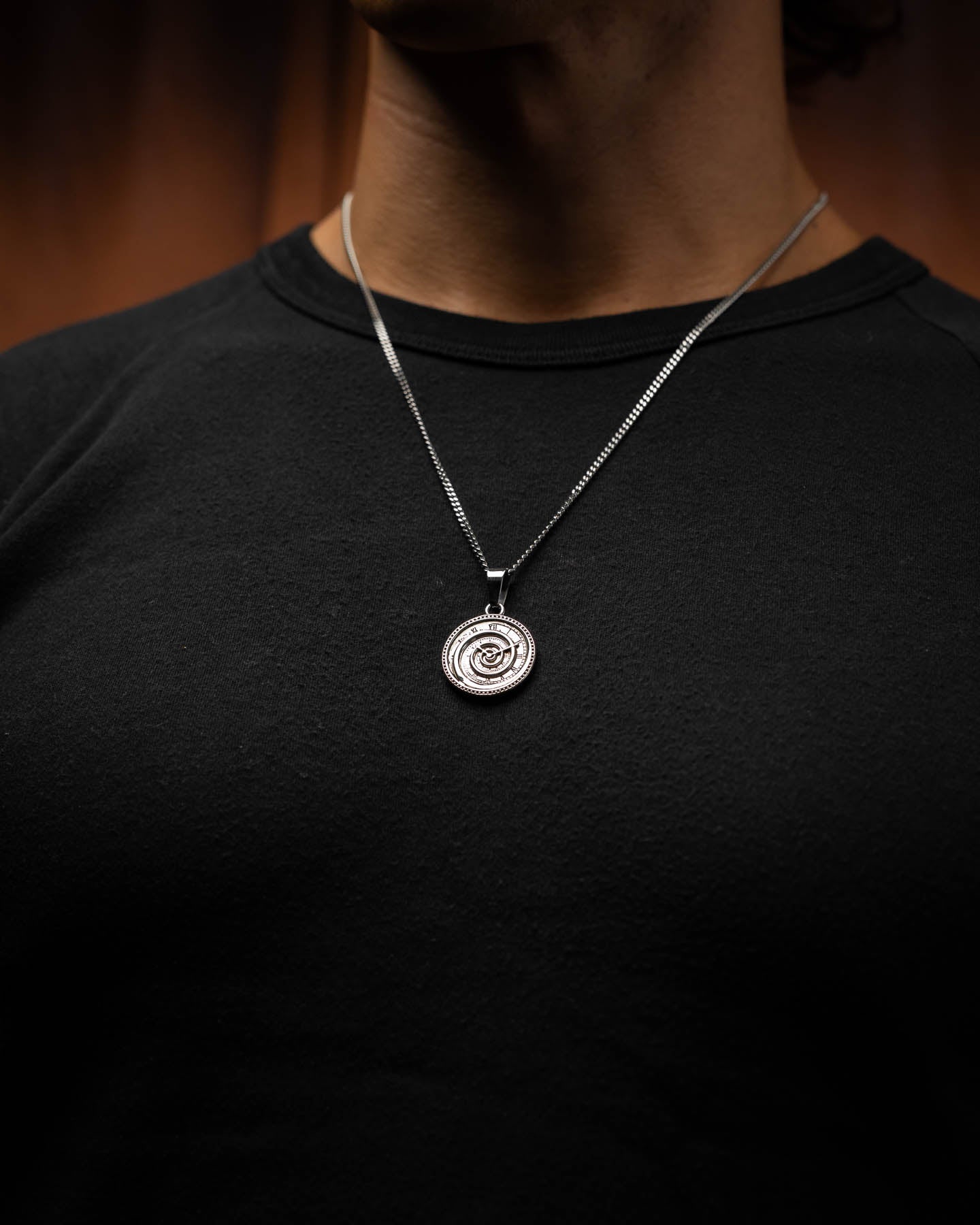 A person wearing a black shirt and the Infinite Clock Pendant Necklace, featuring a silver 316L stainless steel spiral clock design, set against a softly blurred brown background.