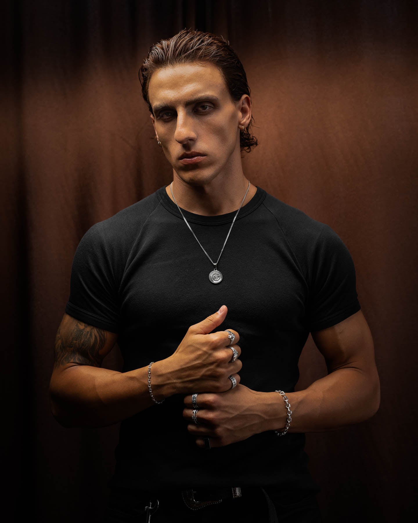 A serious young man with slicked-back hair wears a black t-shirt, 316L stainless steel rings, and the Infinite Clock Pendant Necklace while standing against a dark brown backdrop, gazing confidently at the camera.