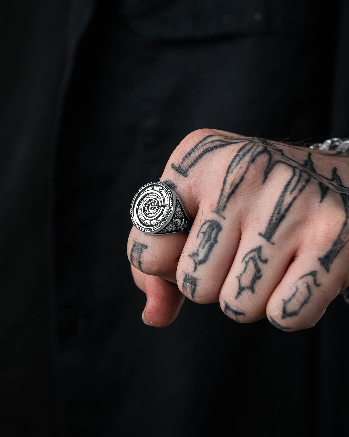 A close-up shows a tattooed hand wearing the Infinite Clock Ring, a large, ornate stainless steel piece with an intricate circular design. The wearer is dressed in black, and the tattoos have bold, dark lines and shapes.