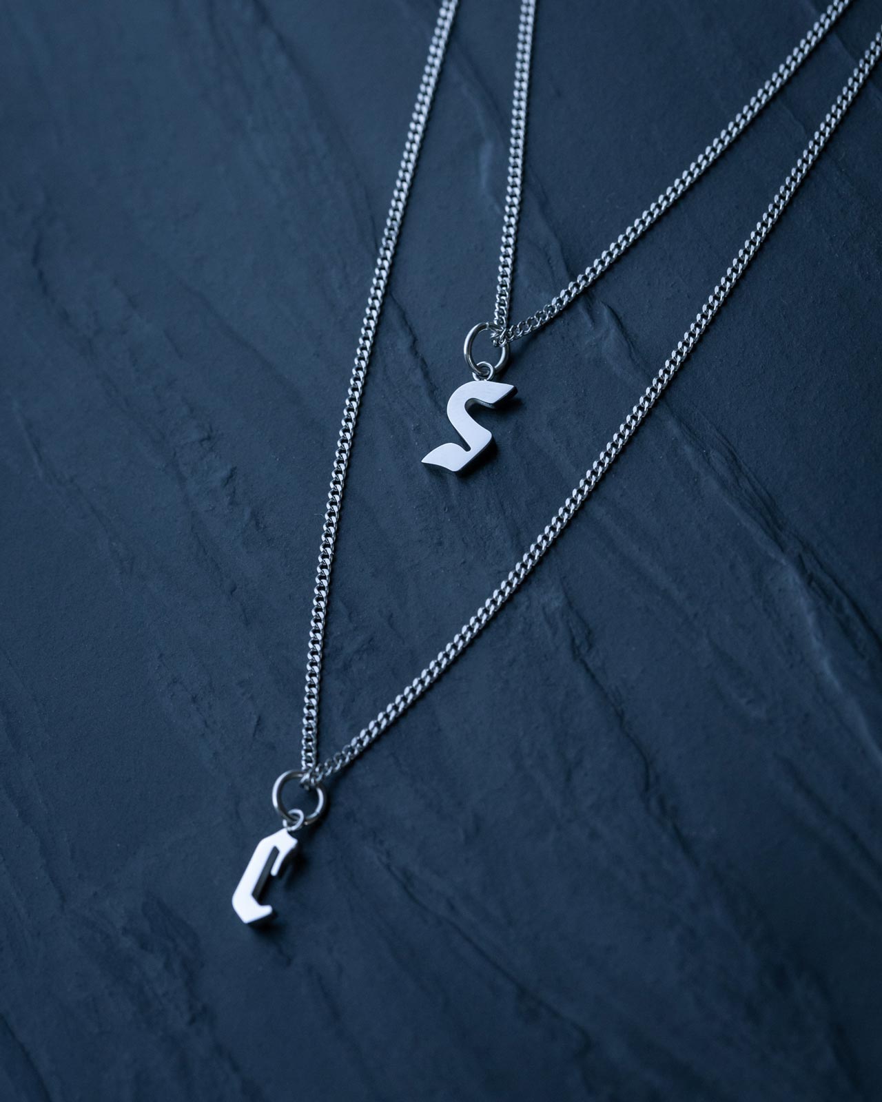 Two silver curb chains with Custom Initial Letter Pendants—S and C—from Statement Collective rest on a dark stone surface, perfect for fans of personalized necklaces.