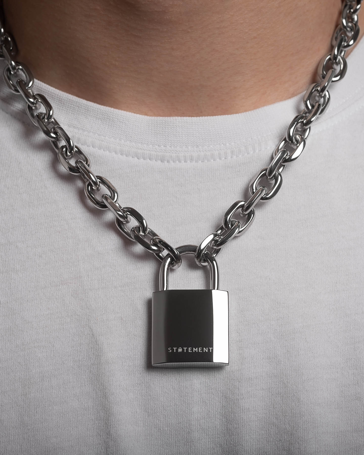 A close-up of a person wearing the Statement Lock & Chain 2.0—a thick silver necklace featuring a large padlock pendant engraved with "STATEMENT," styled over a white t-shirt—ideal for bold jewelry enthusiasts.