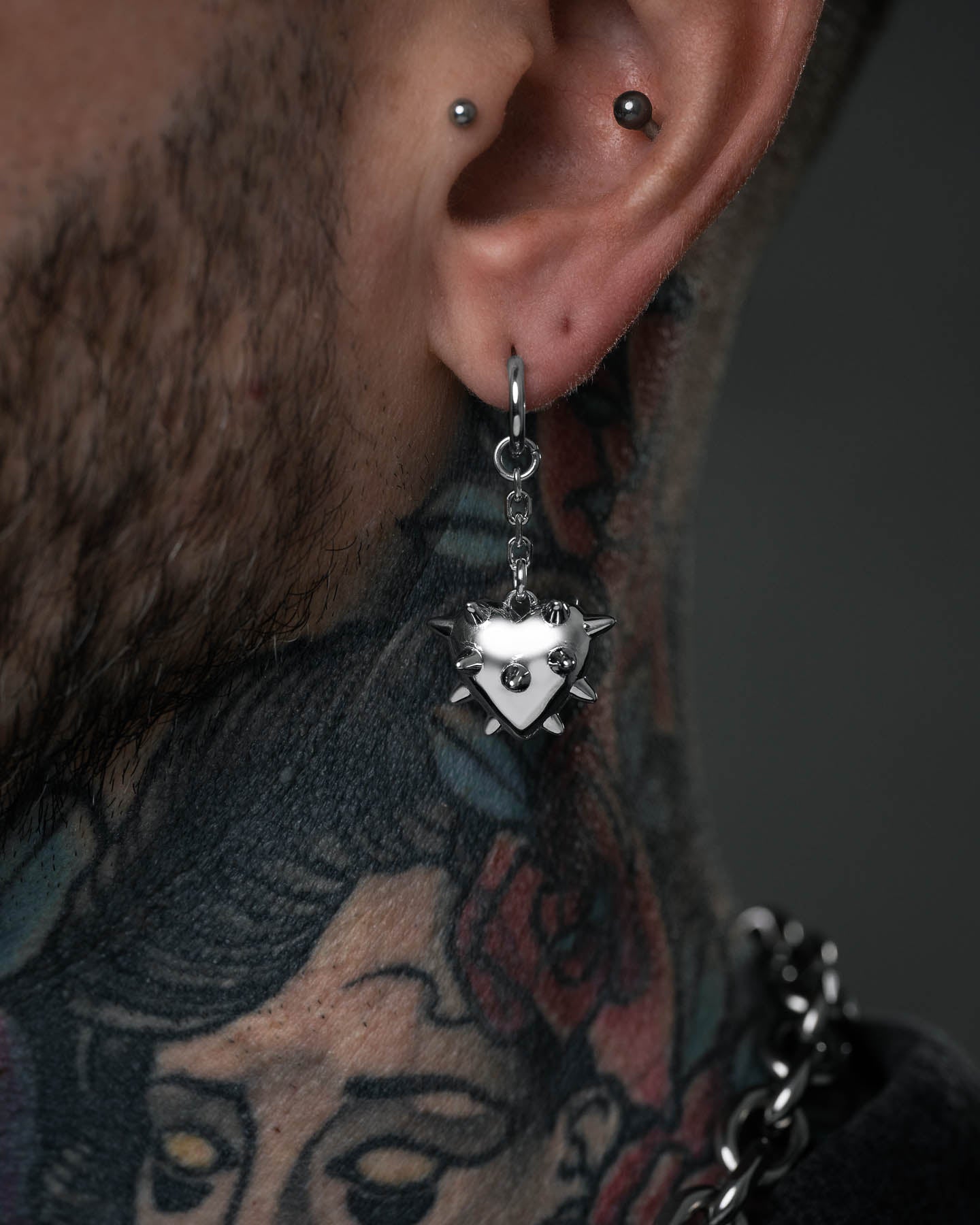 Close-up of a person’s ear with two piercings, wearing the spiked heart-shaped "Love Hurts" Earring. Colorful neck tattoos and a partially visible chunky chain necklace enhance the bold, edgy jewelry style.