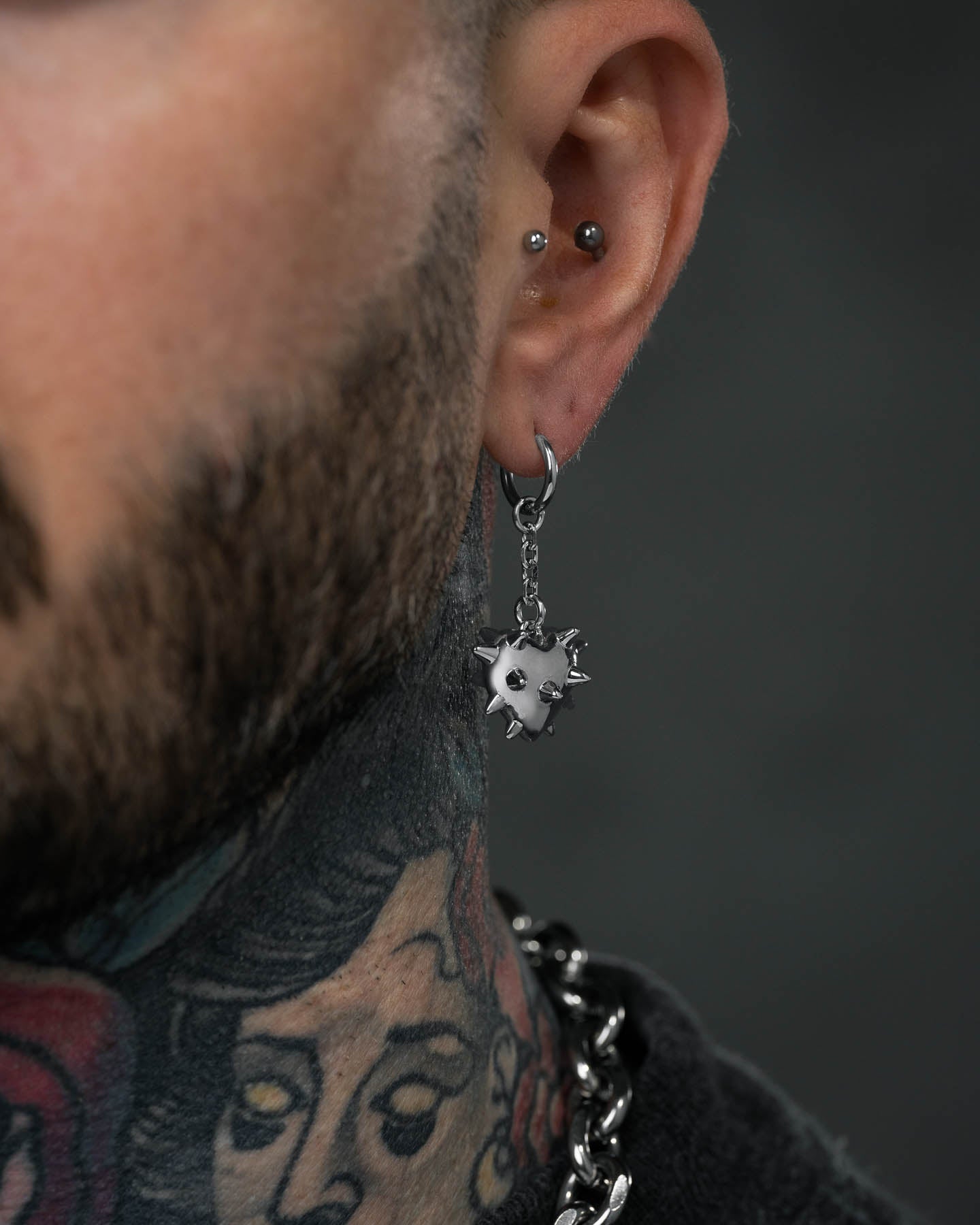Close-up of a bearded person with neck tattoos, wearing multiple piercings including the edgy "Love Hurts" Earring. They also sport a chunky metal chain necklace, all set against a blurred background.