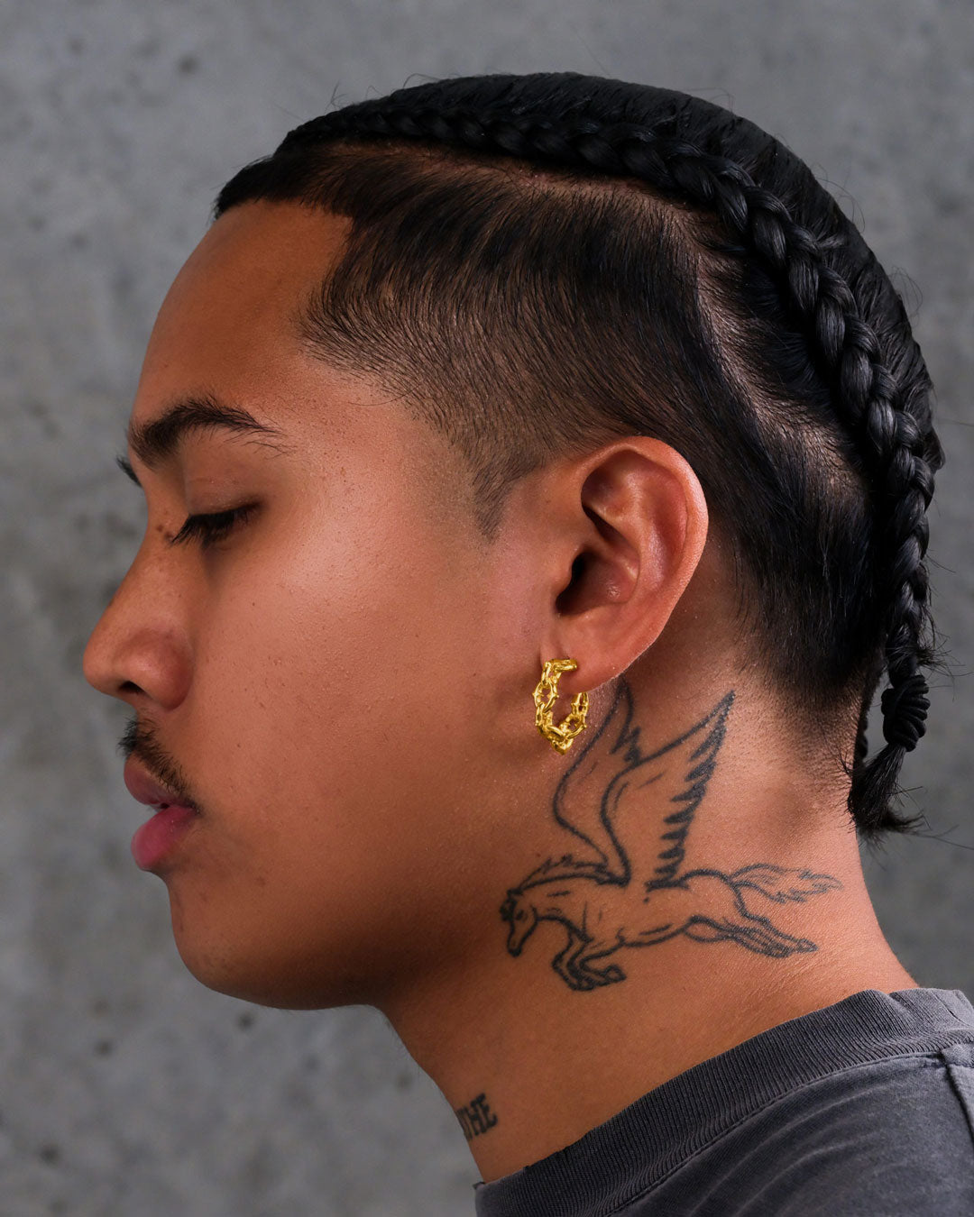 A person with braided hair and a mustache is shown in profile, wearing "The Cathedral" Hoop Earrings (Gold) and displaying a winged horse tattoo on their neck. The background is a plain gray surface.