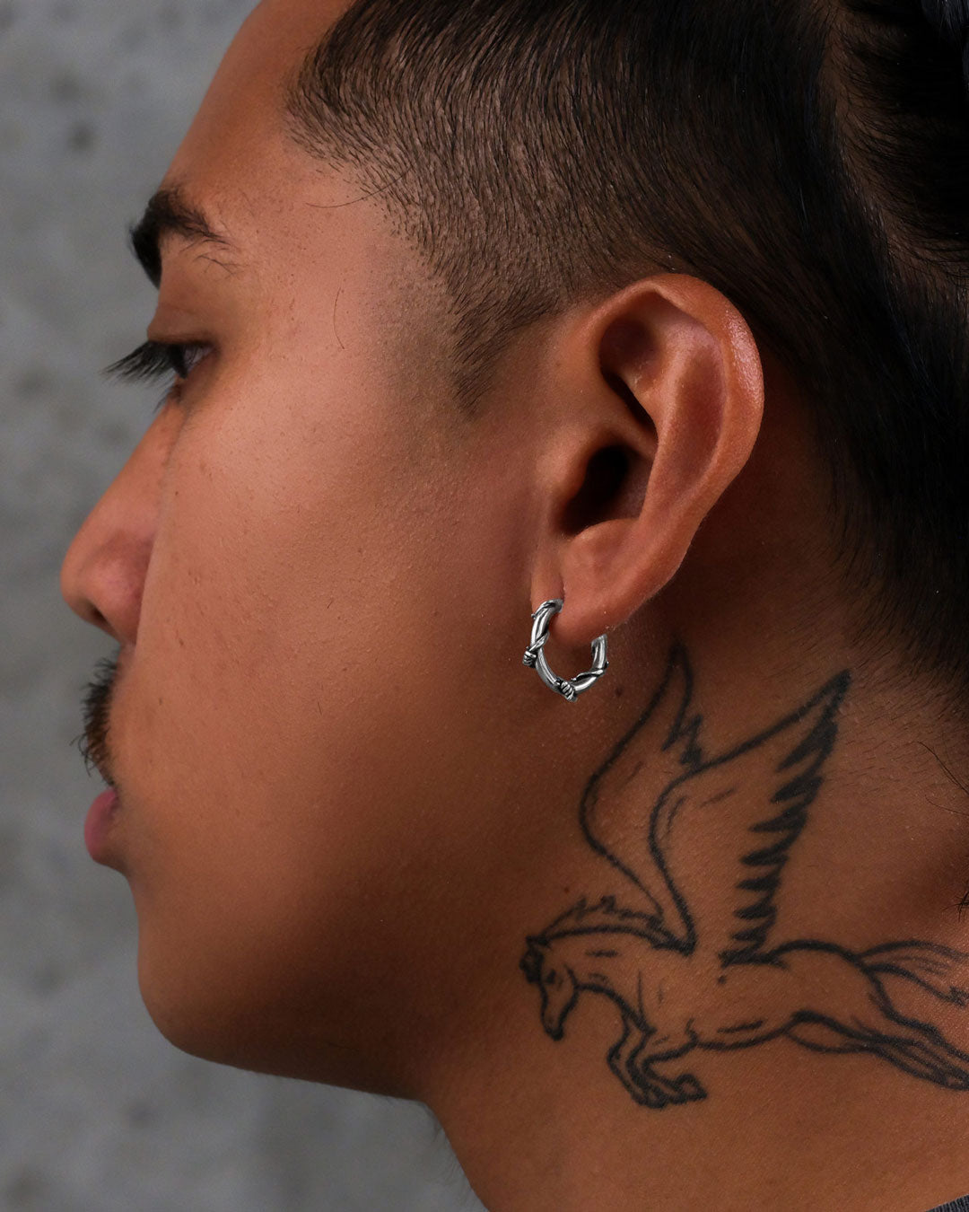 A person with short hair and a mustache wears Barbed Wire Hoop Earrings. A large tattoo of a winged horse, resembling Pegasus, is visible on their neck. The background is a neutral, blurred gray.