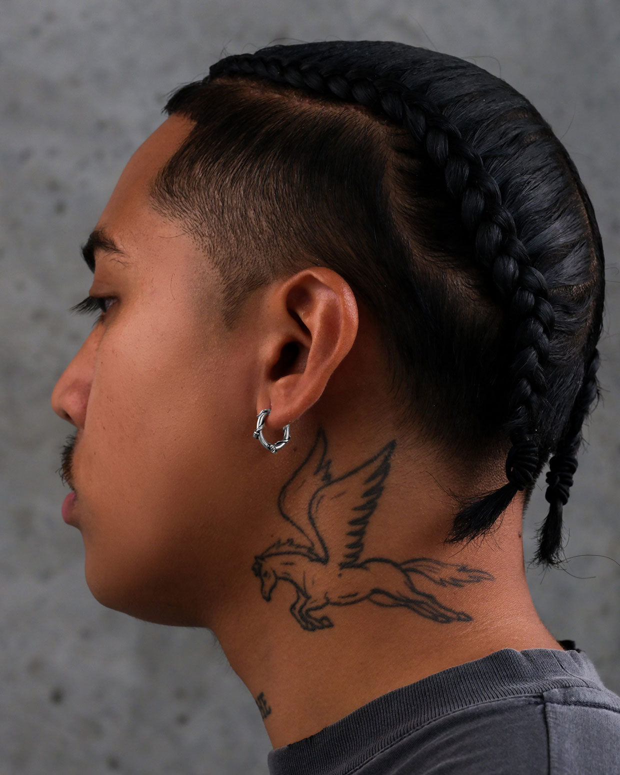 A person with braided hair and a mustache is shown in profile, wearing Barbed Wire Hoop Earrings and displaying a Pegasus tattoo on their neck against a gray textured background.