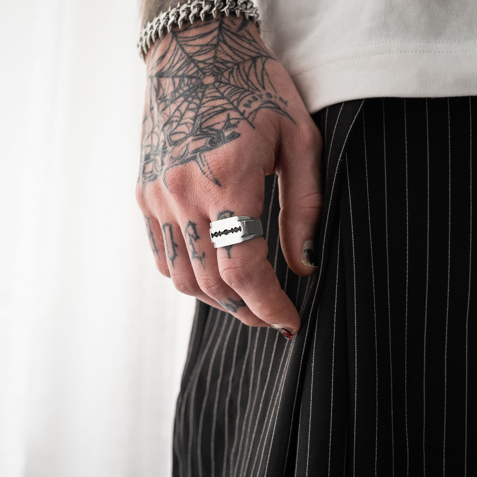 Hand with tattoos wearing a silver razorblade signet ring, against a white background