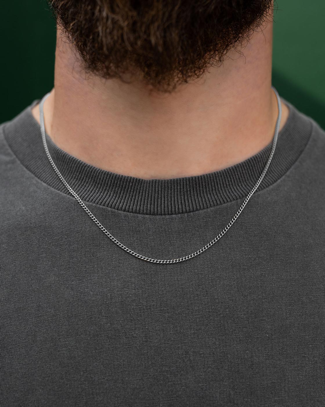 A close-up shows a person wearing Statement Collective's 2.3mm Curb Chain over a dark gray crew neck shirt, with only their neck and upper chest visible. A softly blurred green background highlights the chain's elegance.