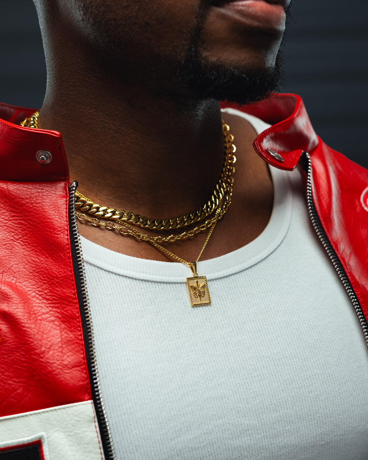 A person wearing a red leather jacket, a white ribbed shirt, and layered gold chains including the Statement Collective Mini Butterfly Tag Pendant in 18k gold. The image is cropped to reveal only the lower half of their face and chest.
