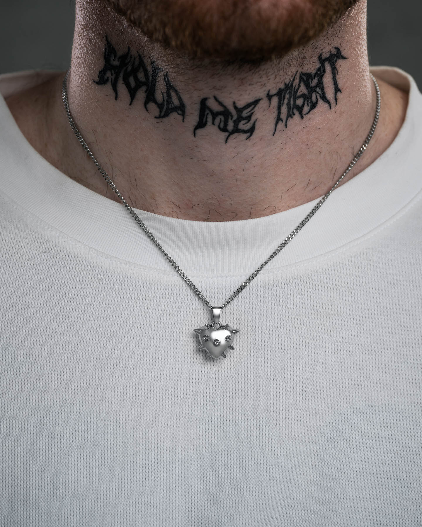 A close-up of a person in a white shirt wearing the Mini "Love Hurts" Spiked Heart Pendant necklace and displaying a bold jagged black neck tattoo reading SHOULD LOVE THYSELF—a striking statement accessory.