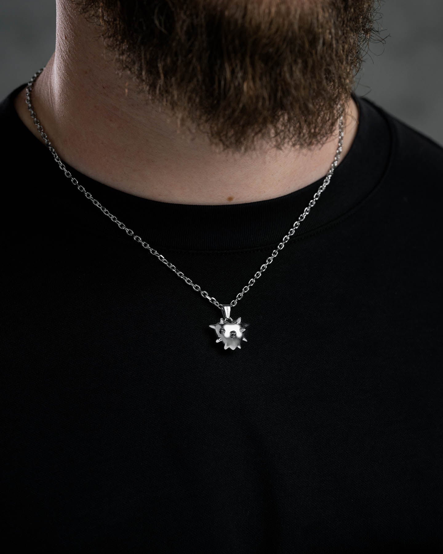 A close-up of a person in a black shirt wearing the Mini "Love Hurts" Spiked Heart Pendant necklace. The dark, blurred background highlights the bold, stainless steel statement jewelry.