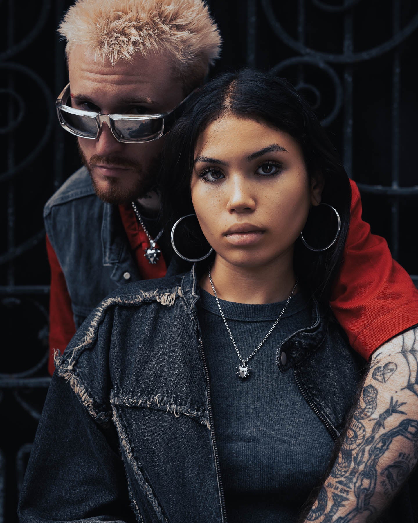 A man with bleached hair and a woman with long dark hair pose by a dark iron gate, both in dark clothes and bold jewelry, including the Mini "Love Hurts" Spiked Heart Pendant. The woman has large hoop earrings and a tattooed arm.
