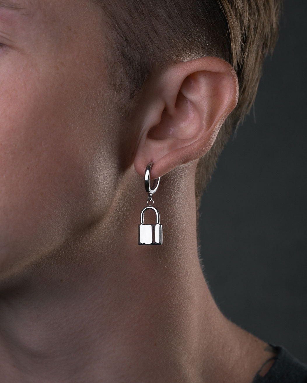 A close-up of a person's ear wearing the Mini Padlock Earring, a minimal men's hoop earring with a small padlock charm. The dark, neutral background highlights this modern accessory.
