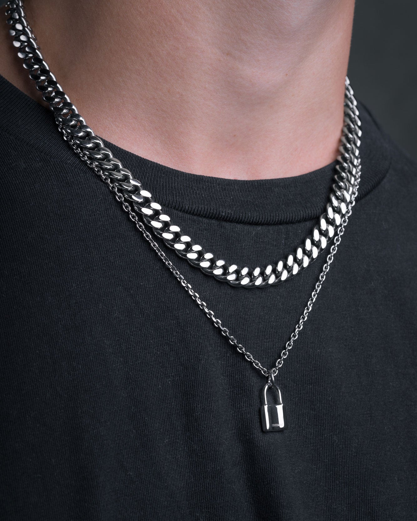 A person wears a black shirt with two silver chains: a thick curb chain and a 2mm chain featuring the Mini Padlock Pendant.