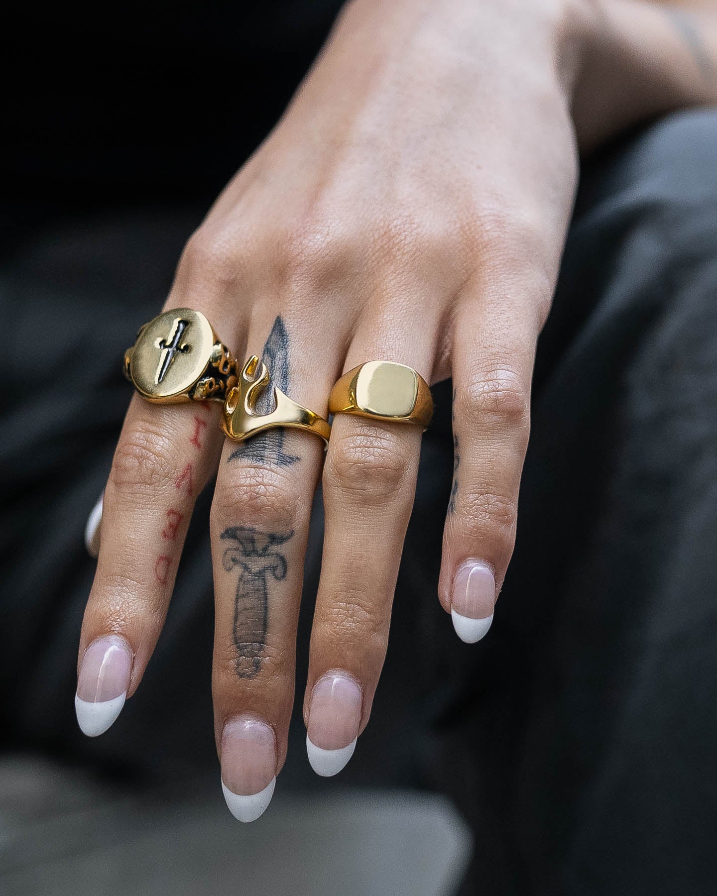 A hand with French-manicured nails, tattoos, and three rings—an 18k gold cross ring, the Minimal Signet Ring (Gold), and a script ring—rests on the knee.