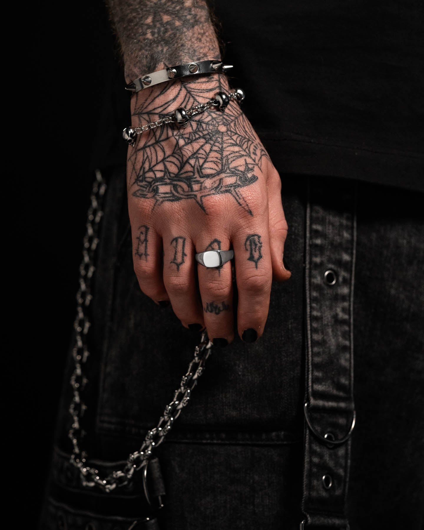 A tattooed hand with black nail polish wears the Minimal Signet Ring and silver bracelets, resting on dark denim jeans with a chain. The tattoos feature a spiderweb and letters across the fingers.