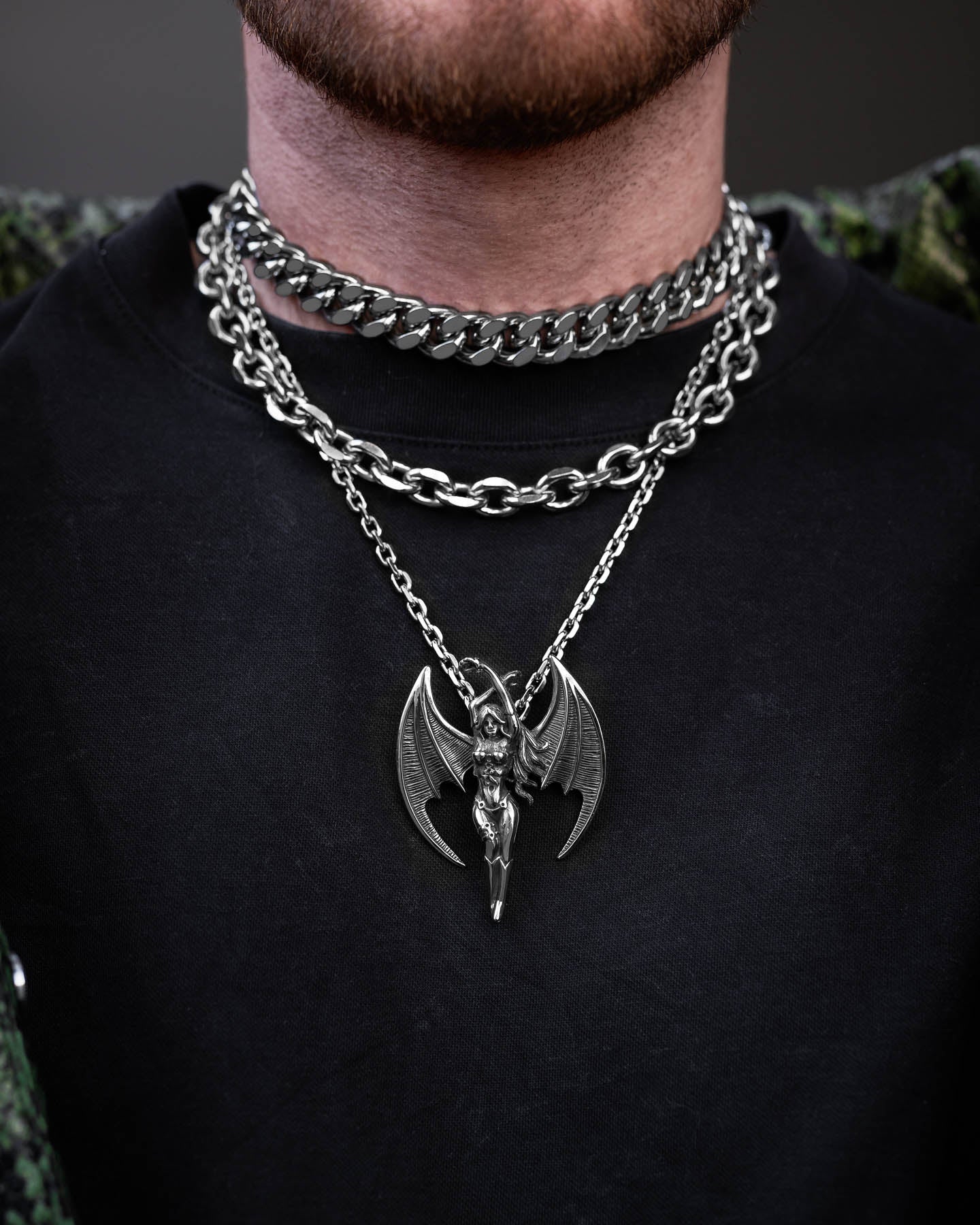 A person wears the Mistress Stack—a thick flat silver chain and a winged demon pendant—over a black shirt and green patterned jacket, with part of their beard visible.