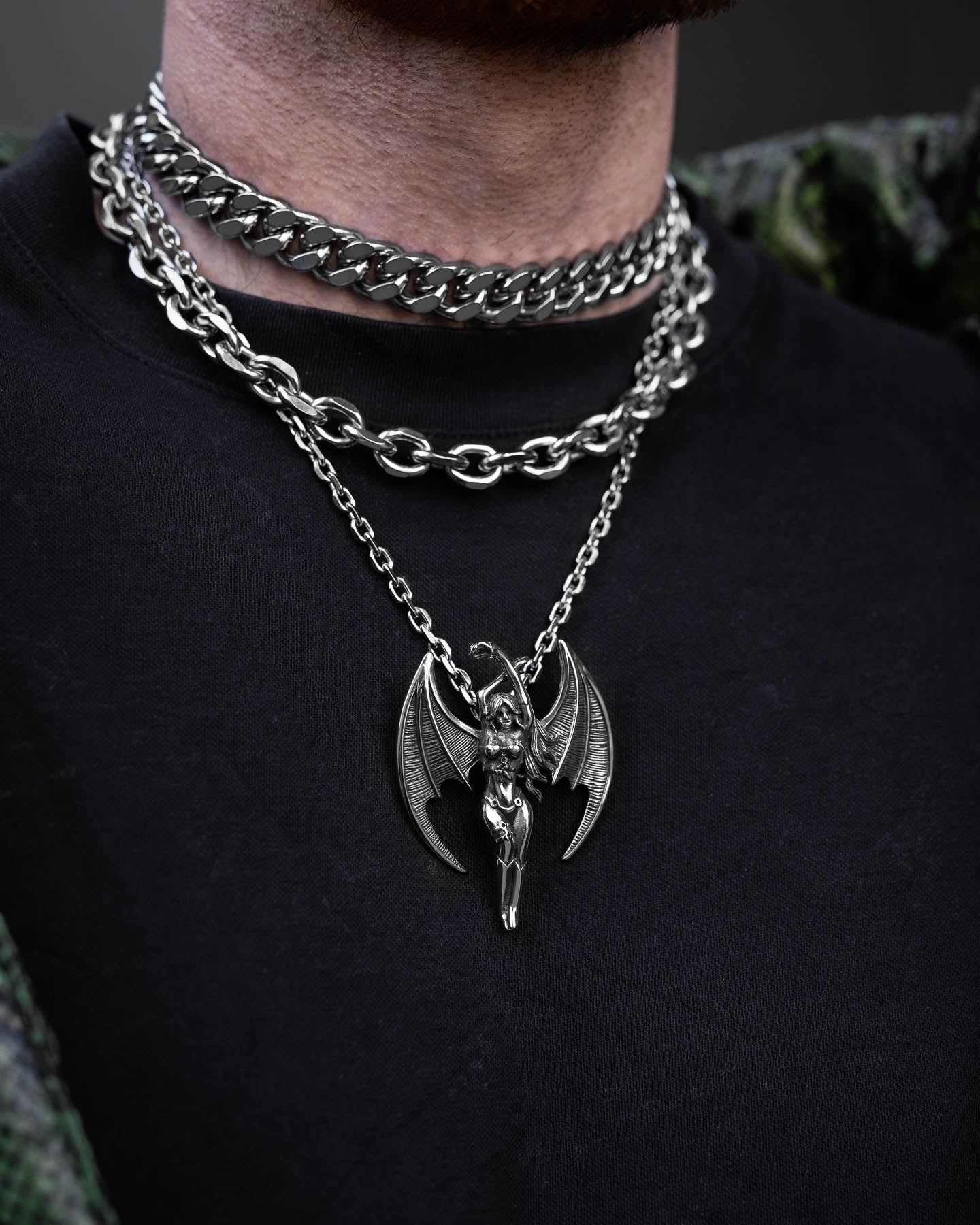 A person in a black shirt showcases the Mistress Stack: two silver chains—a chunky Cuban choker paired with a bold, winged, horned Enchantress pendant.