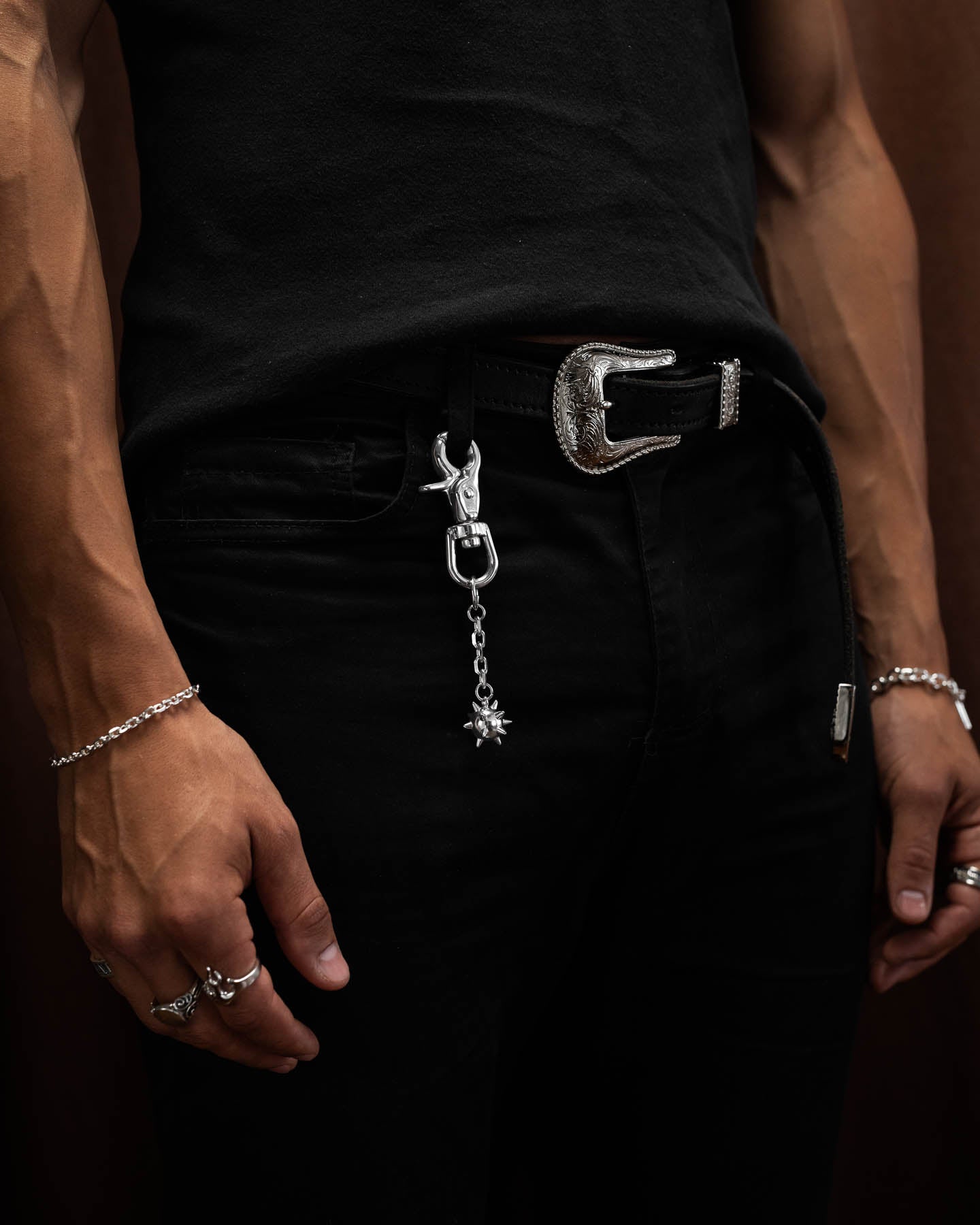 A person wearing a black shirt and pants, accessorized with silver rings, a bracelet, a textured belt with a detailed buckle, and the Morning Star Belt Clip—a 316L stainless steel accessory with decorative charms—hanging from a belt loop.