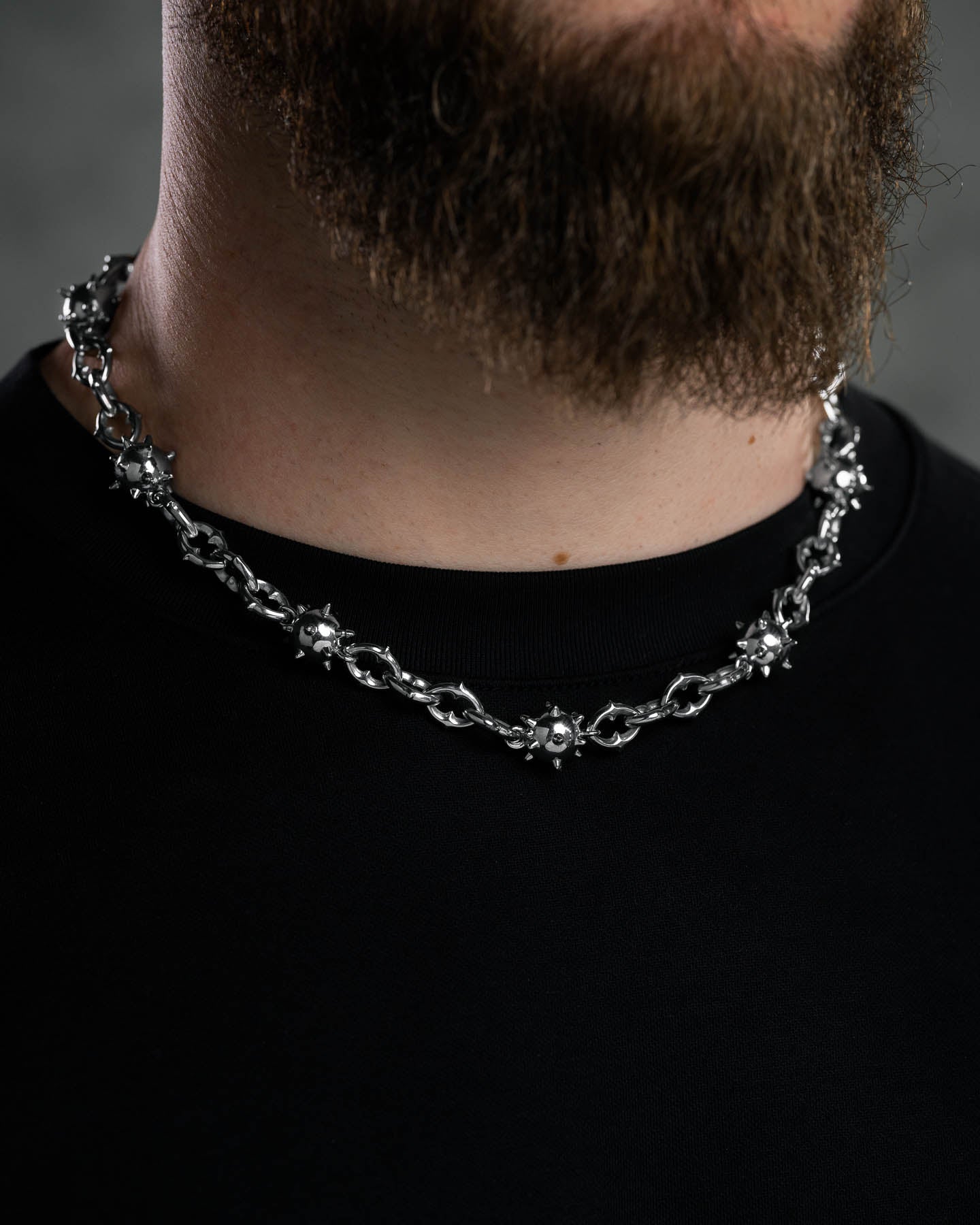 A close-up of a person with a brown beard in a black shirt wearing the "Morning Star" Cathedral Chain, a chunky stainless steel necklace with small spiked ball designs inspired by medieval flail jewelry.