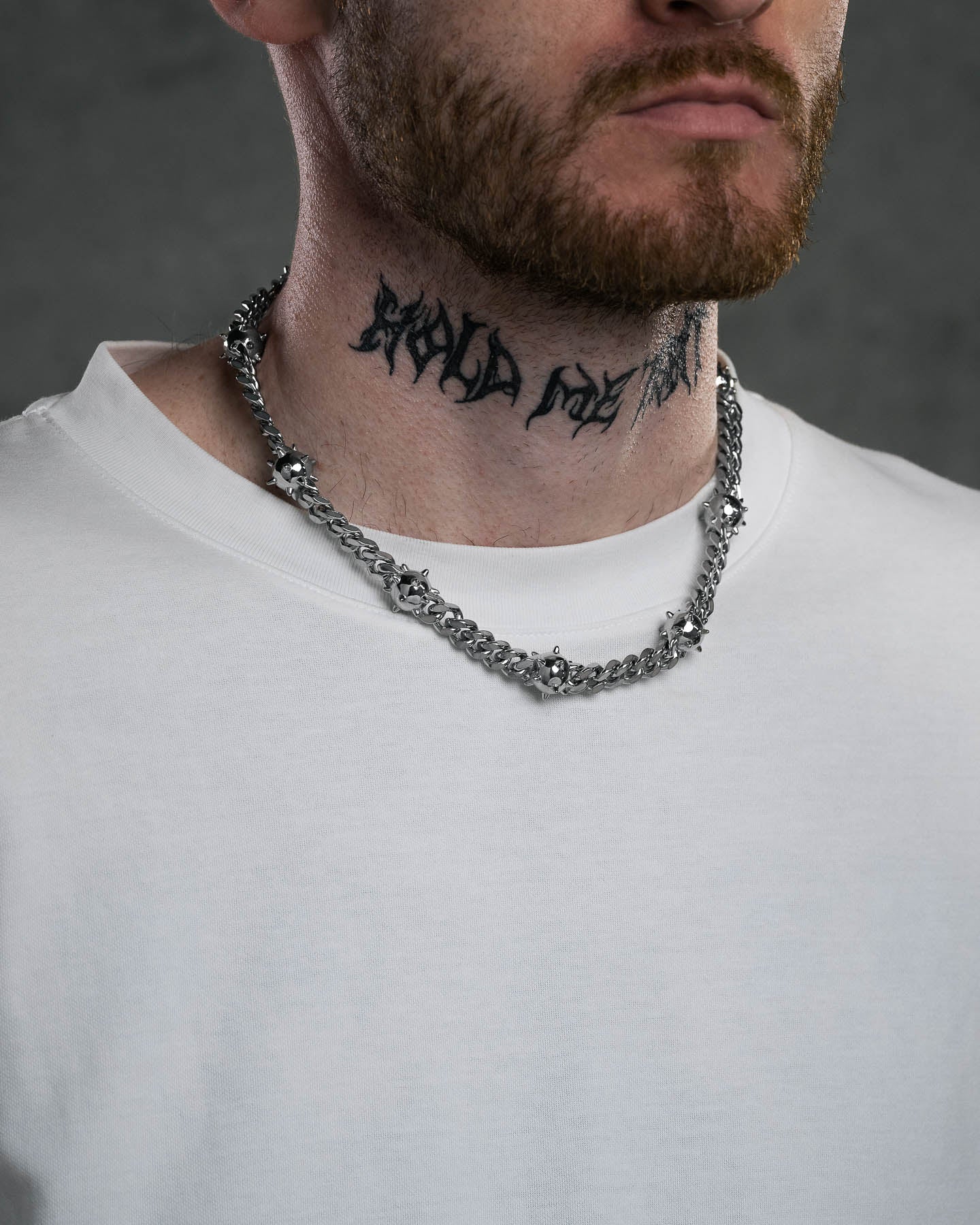 A man with a reddish-brown beard and white shirt wears the "Morning Star" Cuban Chain, a bold stainless steel necklace, while a black tattoo in stylized lettering adorns his neck against a dark, blurred background.