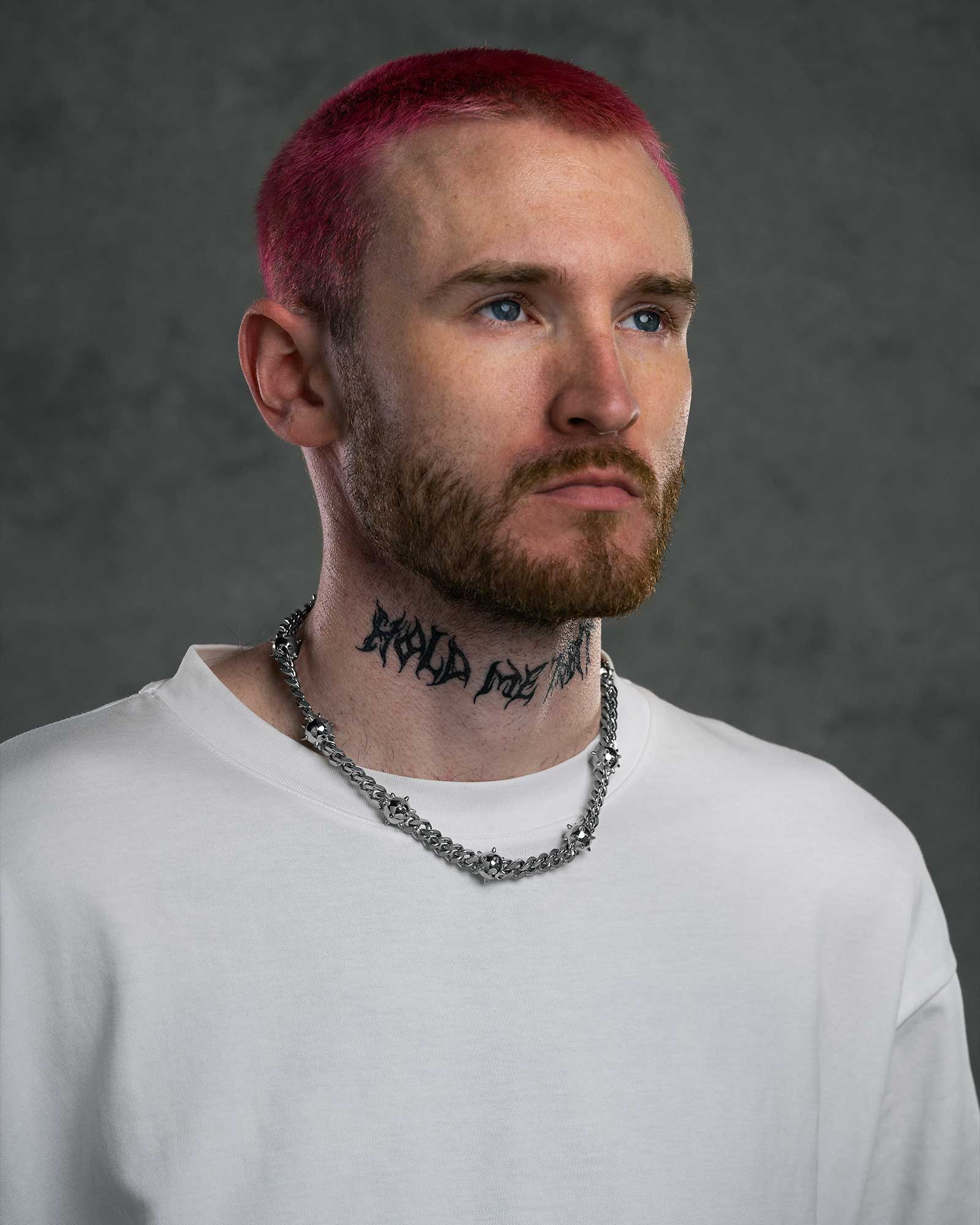 A person with short pink hair, a beard, blue eyes, and a neck tattoo wears a white shirt and the "Morning Star" Cuban Chain. The background is plain dark gray.