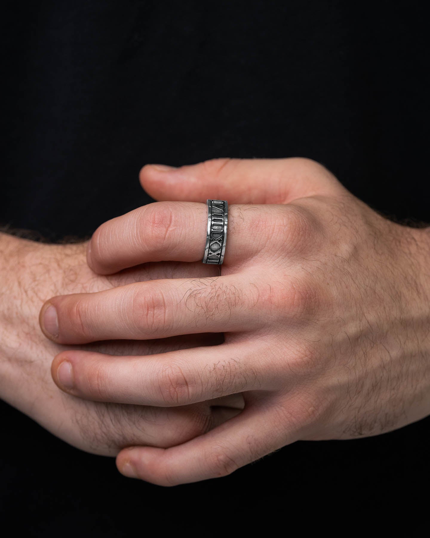 A close-up shows two hands, one resting over the other, wearing the Numerals Ring featuring engraved Roman numerals around the band against a plain black background.