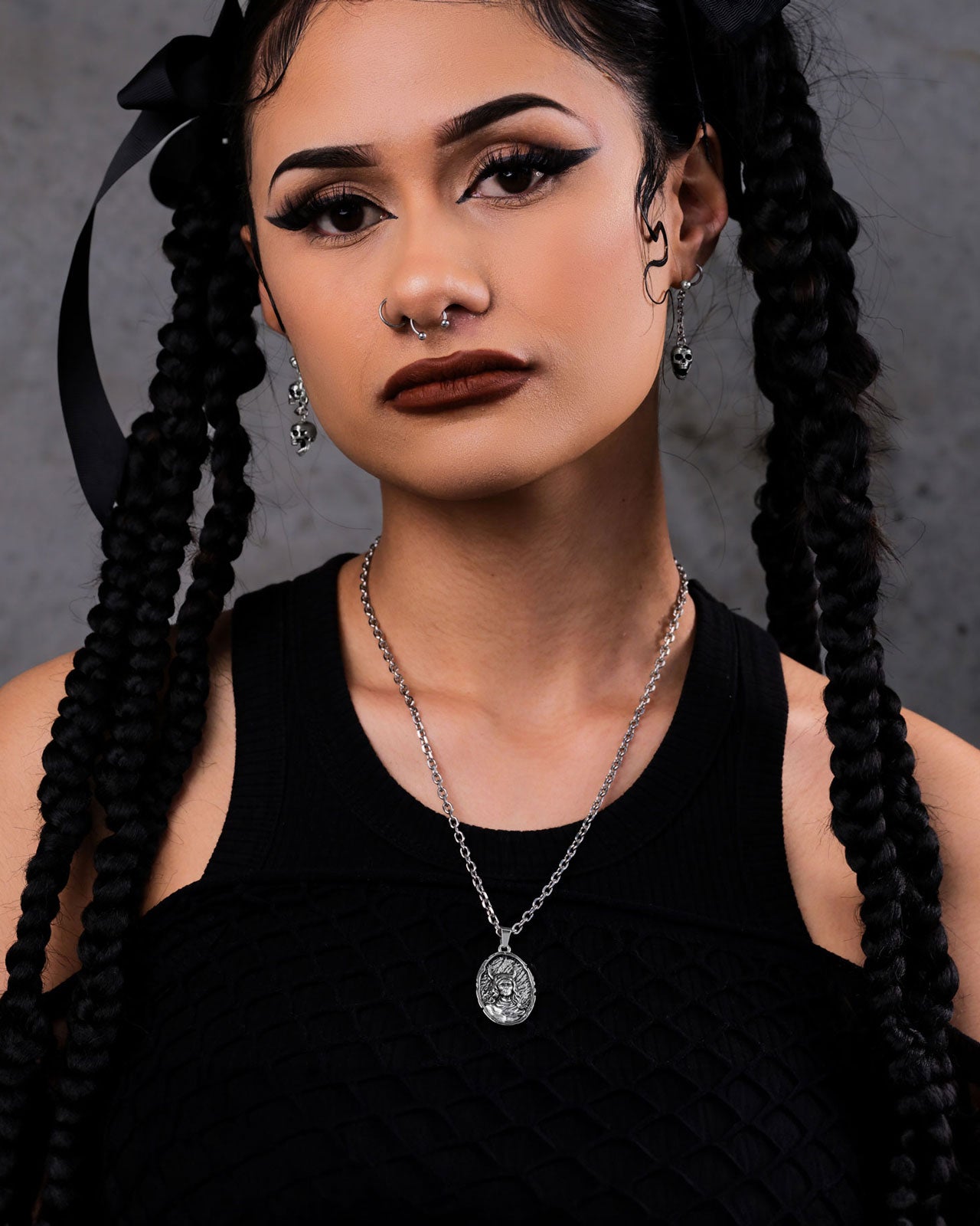 A woman with long braided hair, winged eyeliner, dark lipstick, and a septum piercing wears a black top, silver earrings, and "The Unholy" Pendant gothic stainless steel necklace against a gray background.