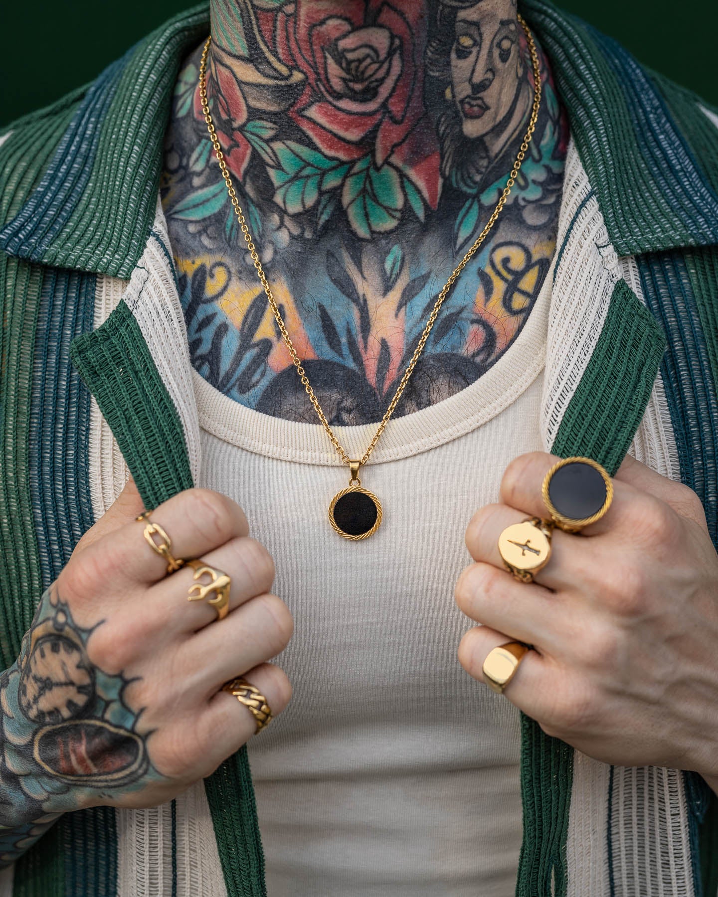 Wearing gold rings and the Onyx Medallion Pendant (Gold), a person with colorful chest and neck tattoos holds open a green and white striped shirt over a cream tank top.