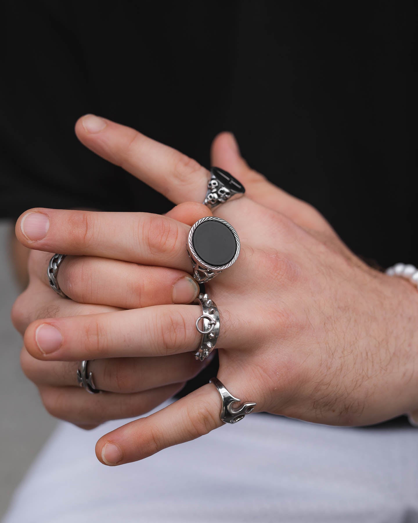 A close-up of two hands wearing multiple silver rings, including the Onyx Sovereign Ring with a large black stone, against a blurred background. The person is dressed in black and white clothing.