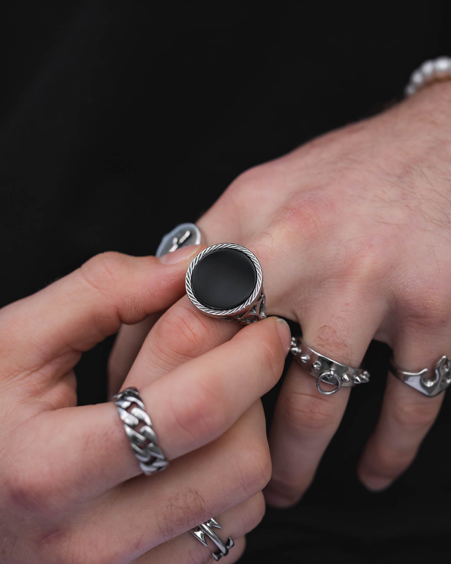 A close-up shows a hand adorned with silver rings, including the Onyx Sovereign Ring—a large, round black agate piece—being adjusted against a dark background that emphasizes its bold, gothic style and intricate design.