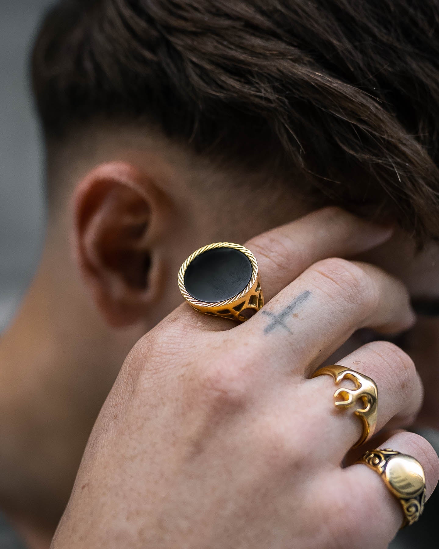 A close-up of a hand with gold rings, including the Onyx Sovereign Ring (Gold), touching their temple. A small cross tattoo is visible on their finger, and they have short brown hair.