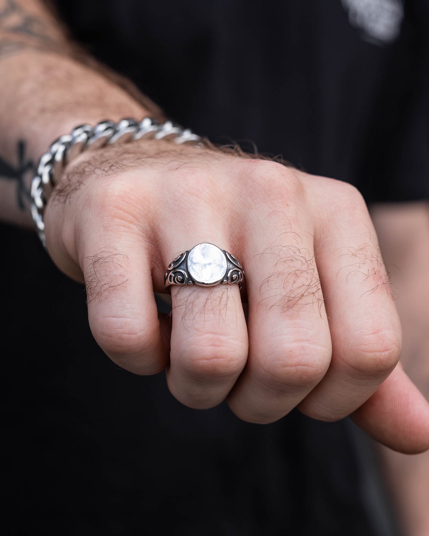 A close-up of a person’s hand wearing the Mini Oval Scroll Signet Ring, featuring a white stone and ornate details, paired with a thick silver chain bracelet. The person is dressed in black.