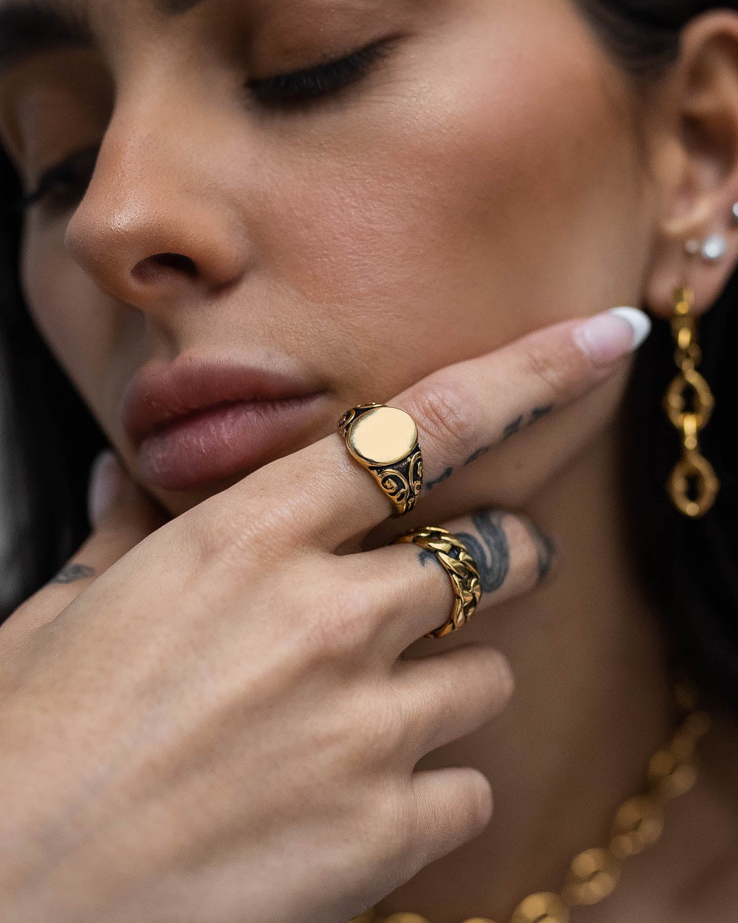 A close-up of a person with long dark hair wearing the Mini Oval Scroll Signet Ring (Gold), a gold necklace, and gold earrings. Their manicured nails with white tips and tattooed finger highlight the perfectly styled waterproof jewelry.