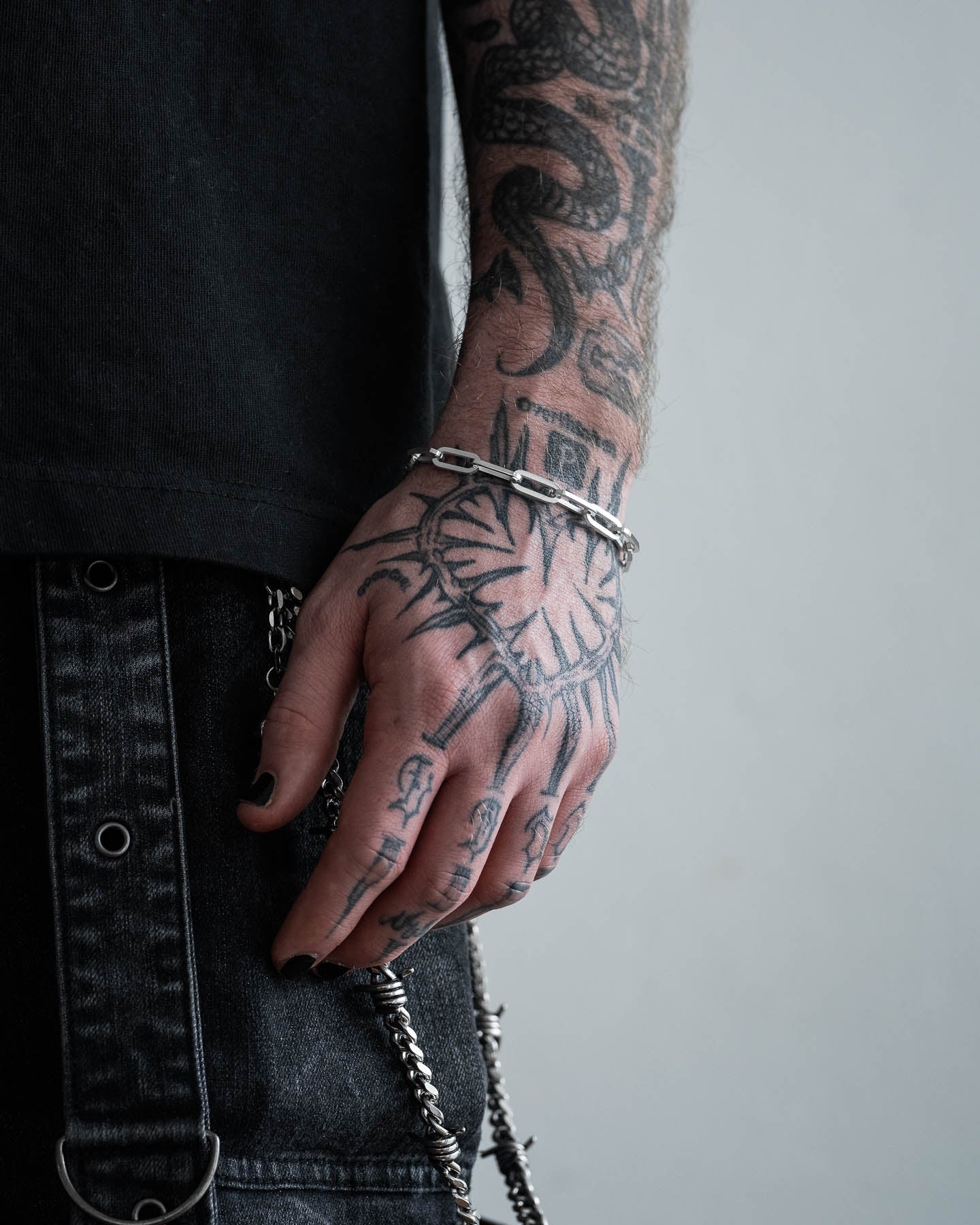 A close-up of a tattooed hand with black nails wearing the non tarnish, waterproof Paperclip Bracelet, paired with a black shirt and dark denim pants detailed with metal chains.