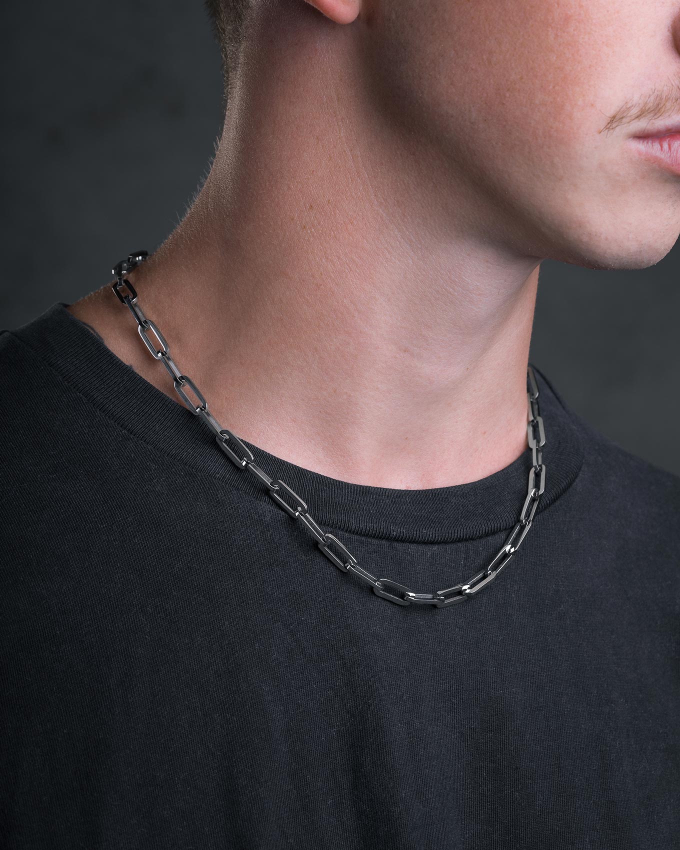 A close-up of a person wearing a black T-shirt and the Statement Collective Paperclip Chain men's necklace, with only the lower part of the face and neck visible.
